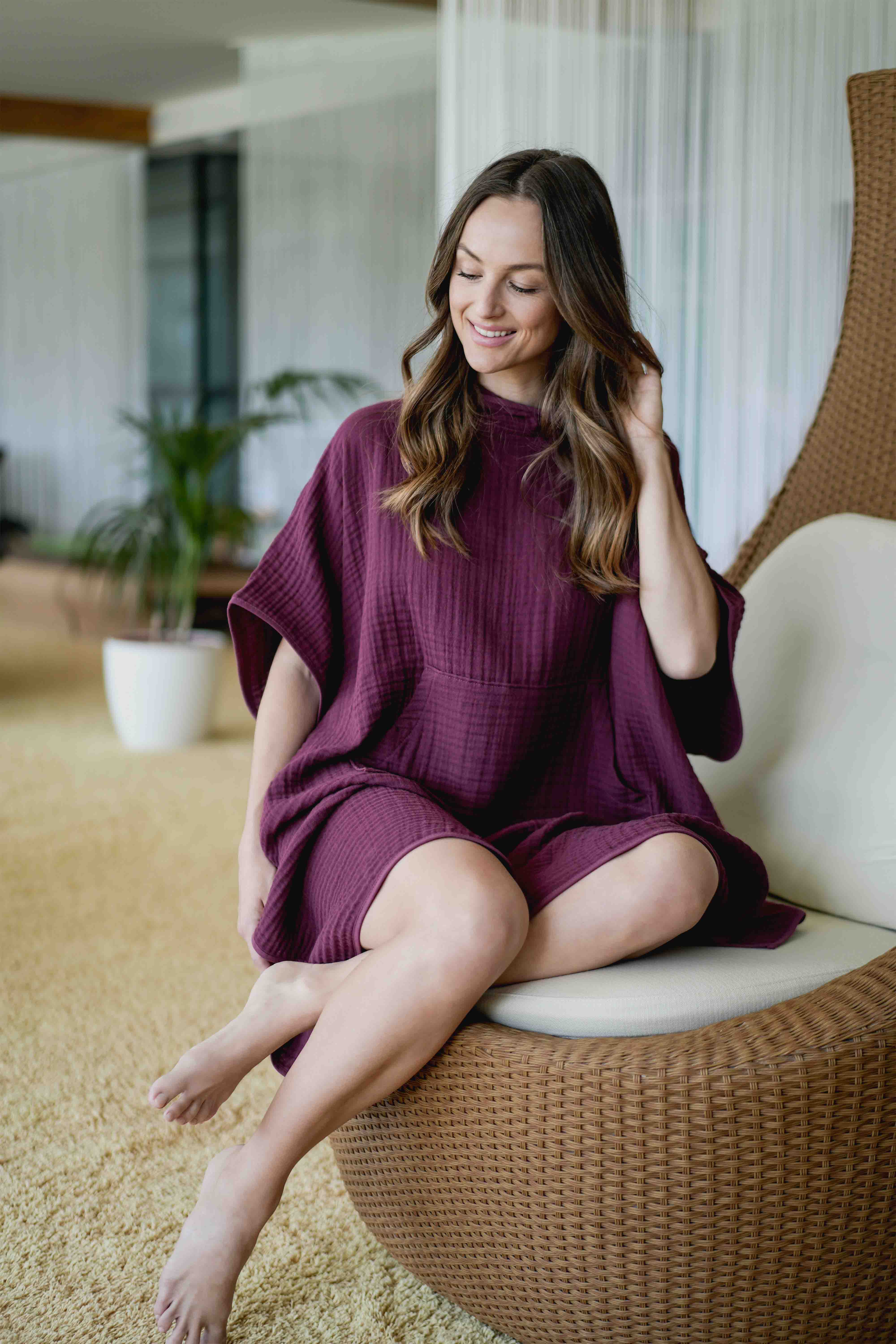 Frau trägt bordeauxfarbenen VOSSEN Spa-Poncho aus Waffelpiqué und entspannt in moderner Wellnesslounge – stilvolle Spa-Mode für pure Erholung.