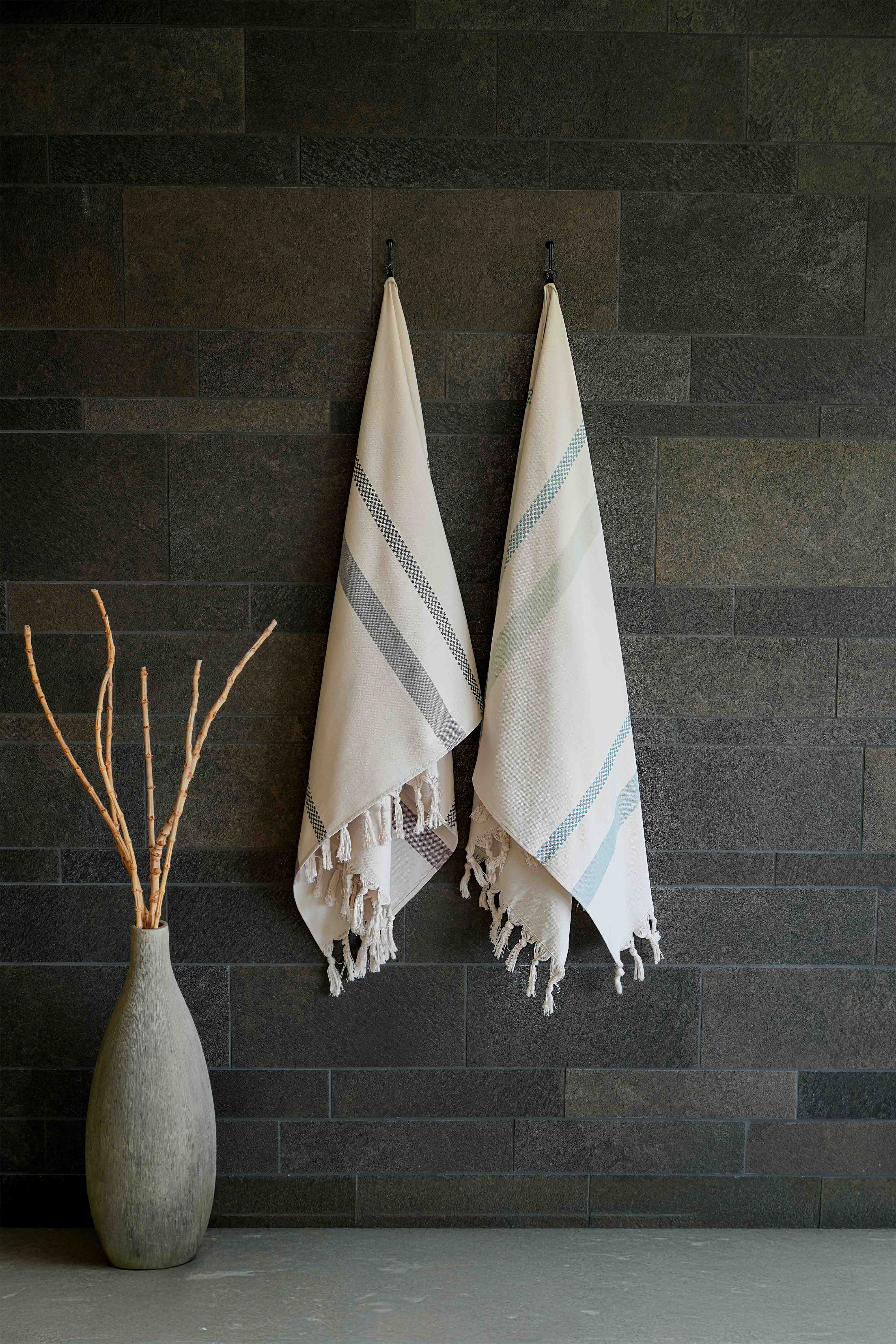 Zwei helle Hamamtücher mit Streifen und Fransen von VOSSEN an dunkler Steinwand – stilvolle Spa- und Wellness-Dekoration mit natürlicher Eleganz.
