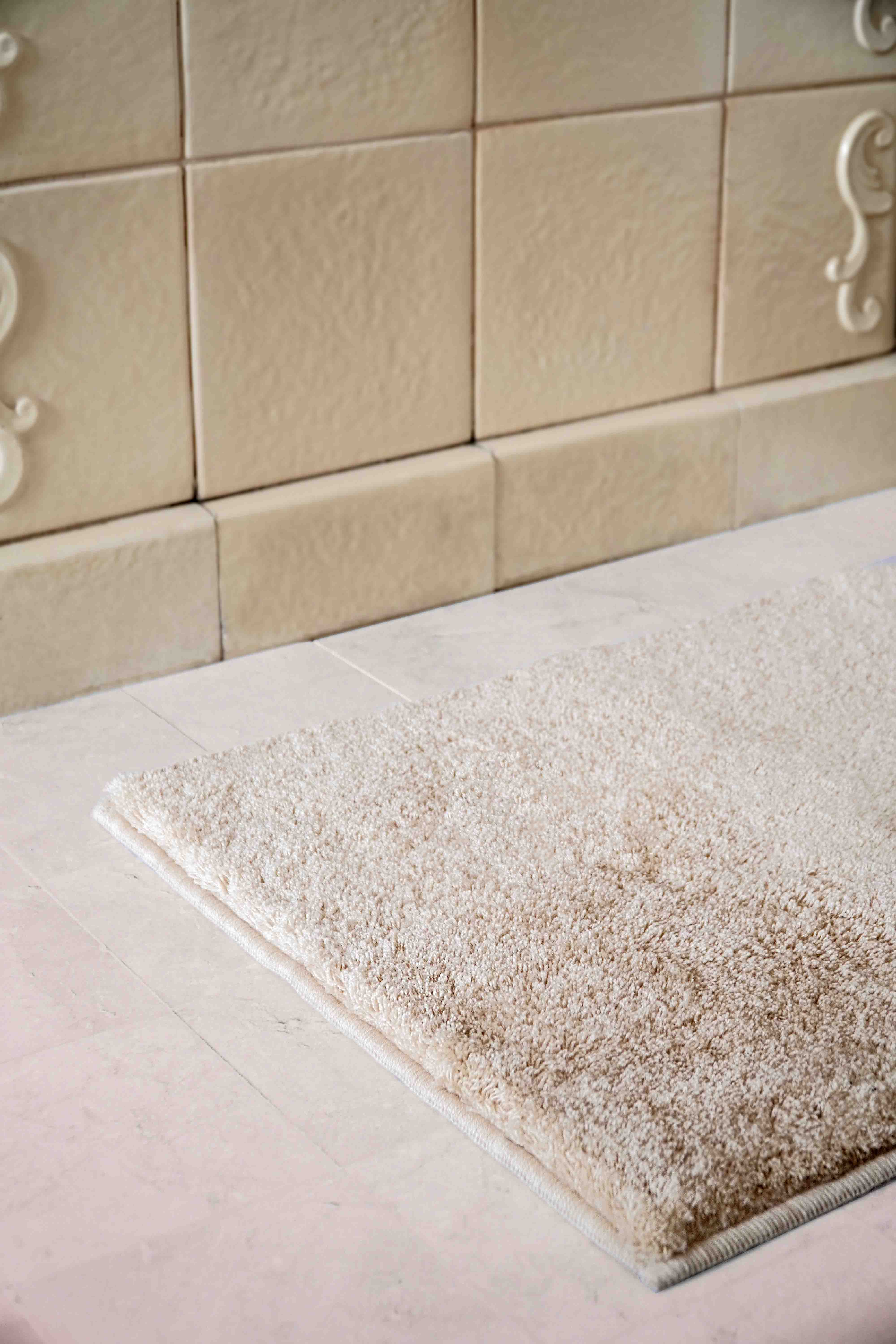 Weicher cremefarbener VOSSEN Badteppich vor beiger Kachelwand – luxuriöser Badezimmerteppich für Wärme und Komfort im modernen Bad.