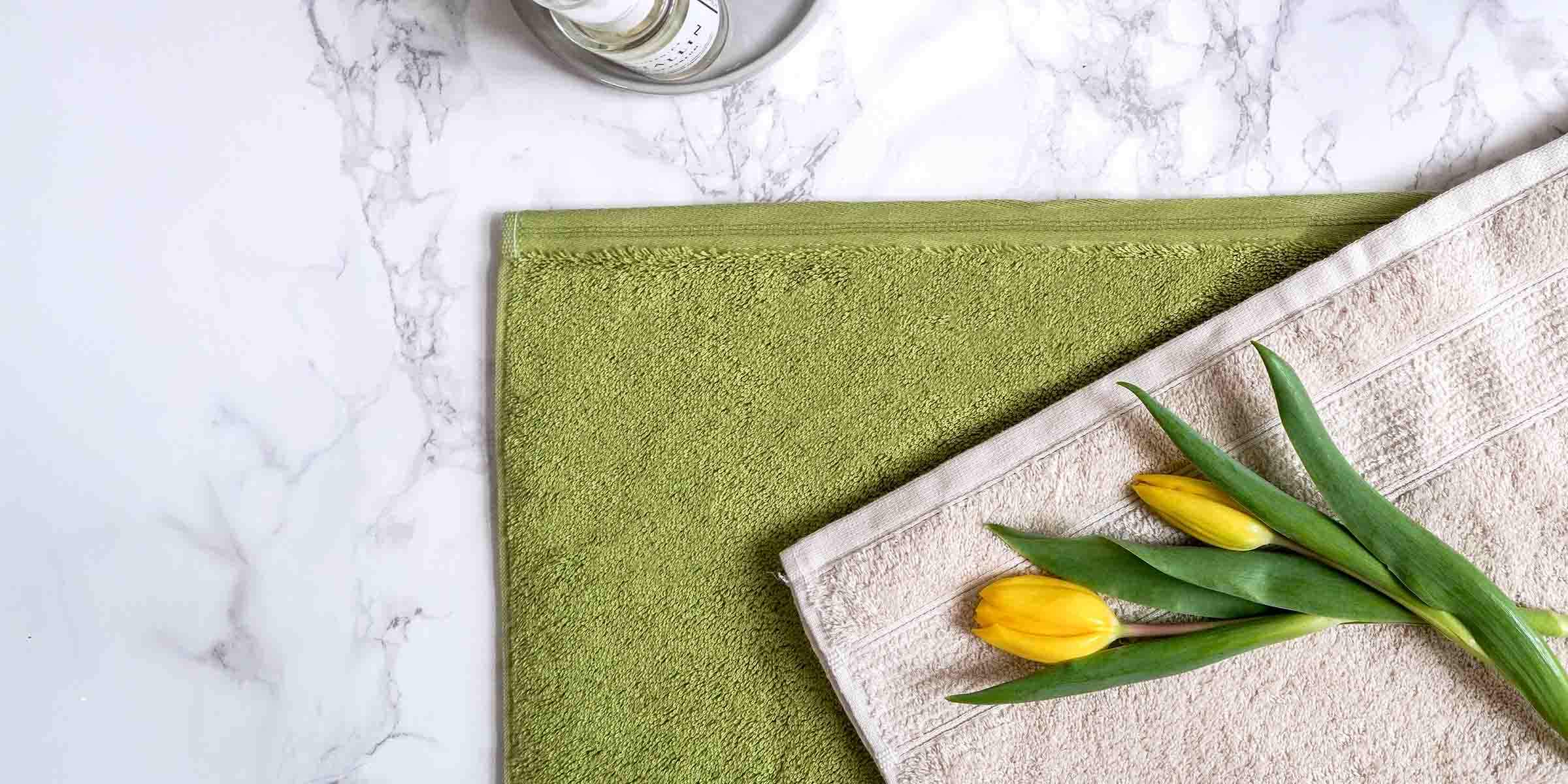 Grünes und beiges Handtuch auf weißem Marmorboden, darauf zwei gelbe Tulpen mit grünen Blättern. Oben links ein Glasgefäß.