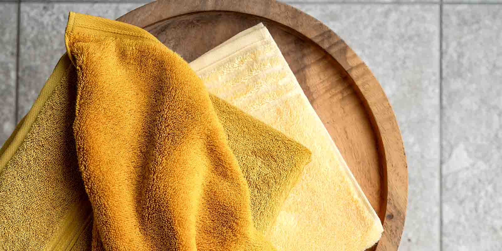 Drei Handtücher in Gelb- und Goldtönen liegen auf Holzfläche, eines ist über die anderen gelegt. Im Hintergrund Fliesenboden.