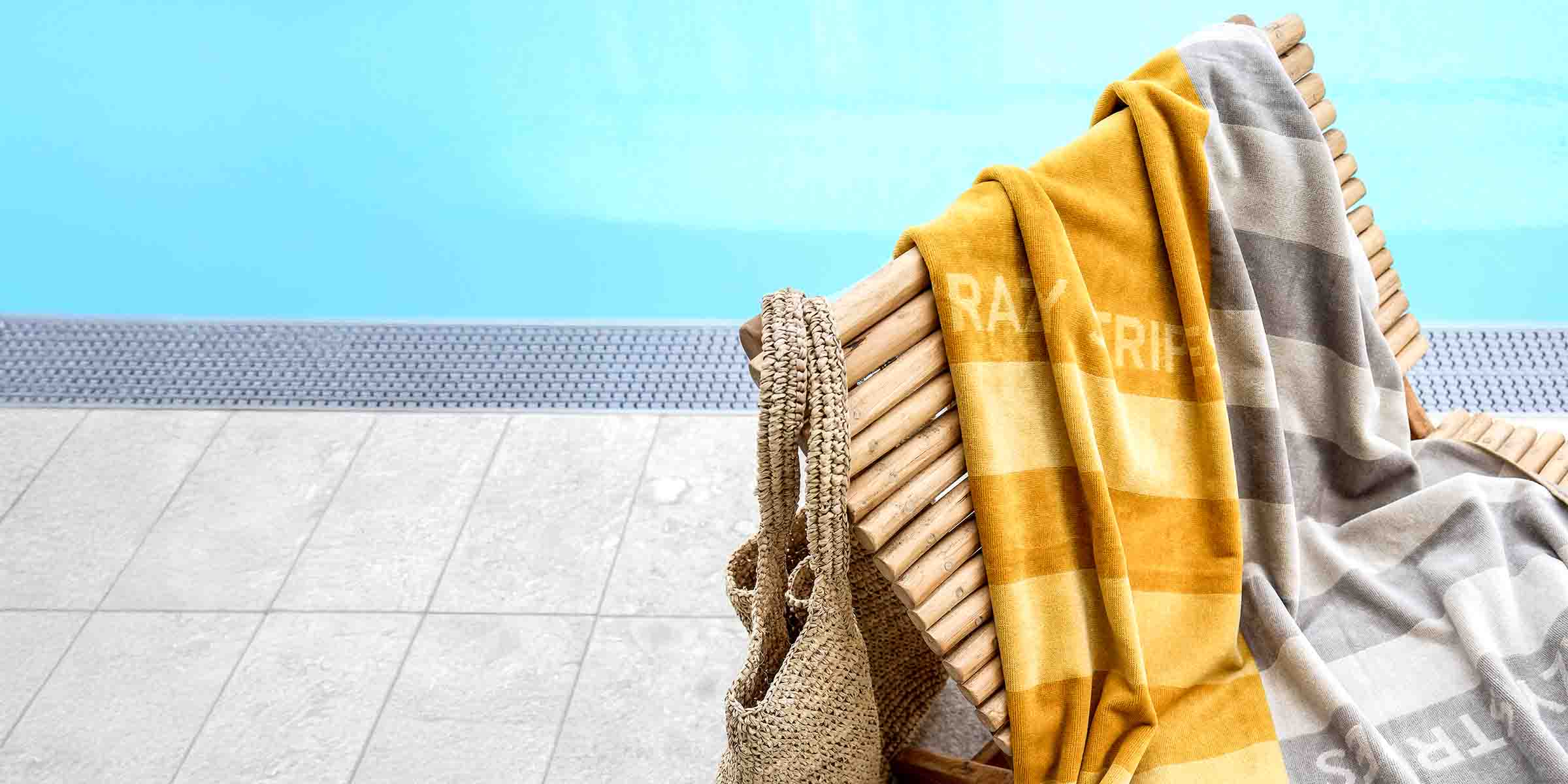 Poolszene mit Holzliege, darauf ein gestreiftes Handtuch in Gelb, Grau und Weiß. Eine geflochtene Tasche hängt an der Liege. Im Hintergrund blauer Pool und Fliesenboden.