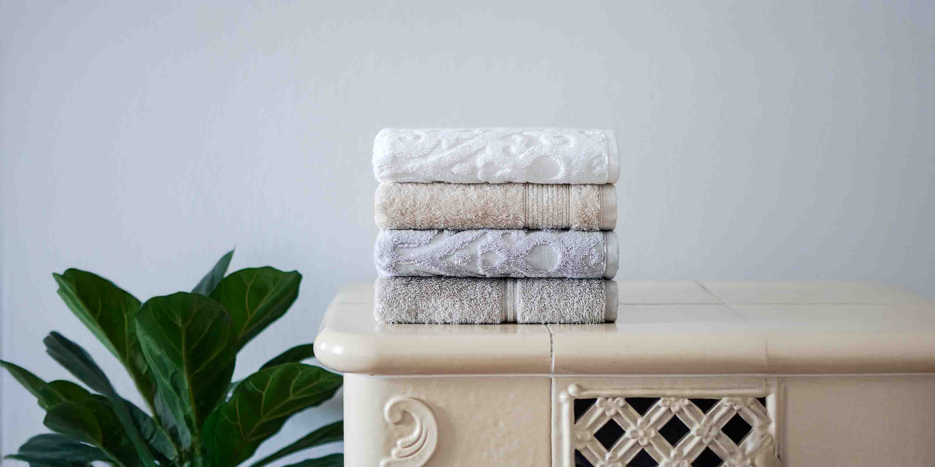 Vier gefaltete Handtücher in Weiß, Beige und Grautönen liegen gestapelt auf beigem Möbelstück mit Deko. Links daneben eine grüne Pflanze mit großen Blättern.