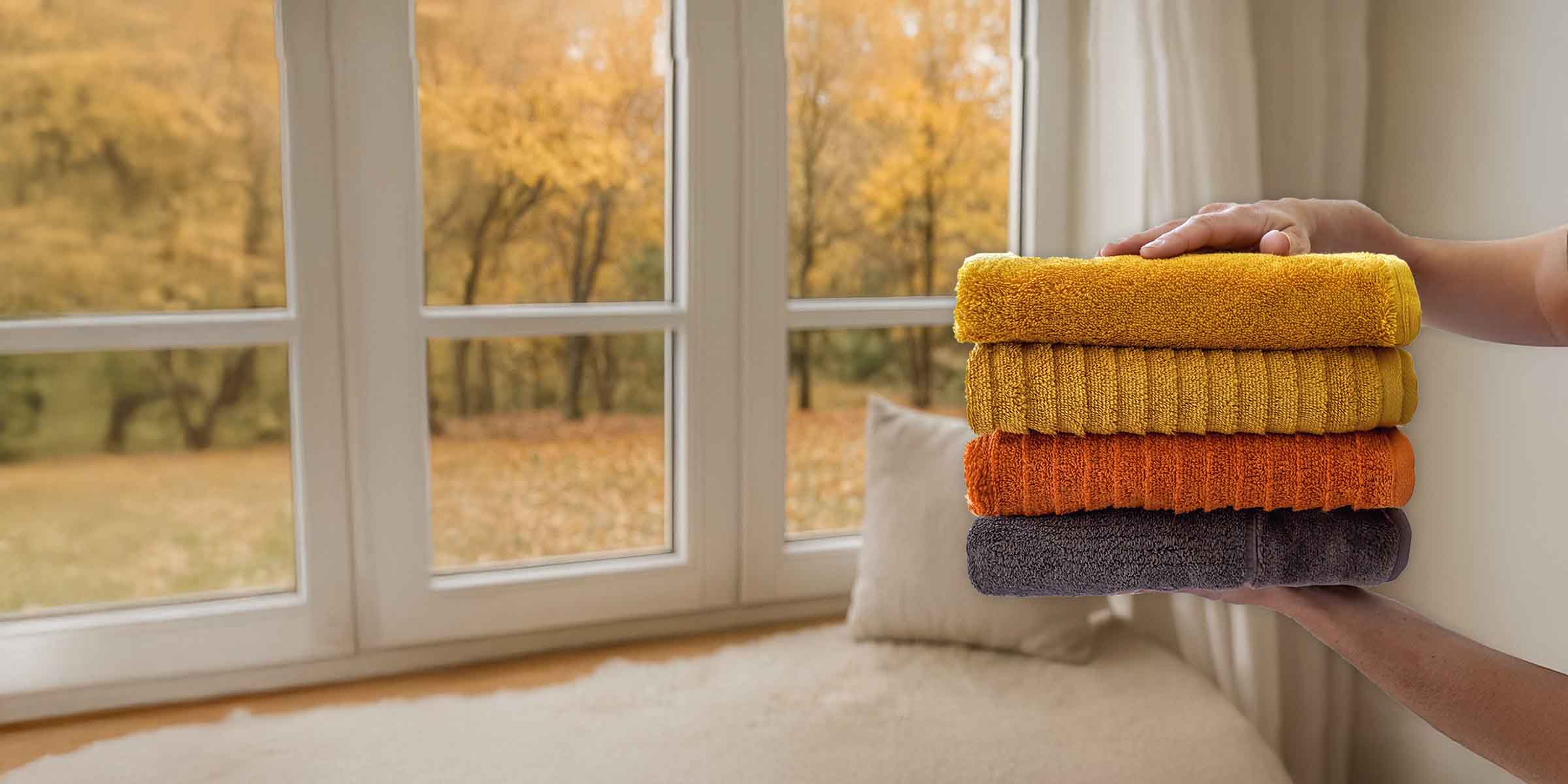 Weiche Handtücher in warmen Herbstfarben liegen vor einem großen Fenster mit Blick auf goldene Bäume.