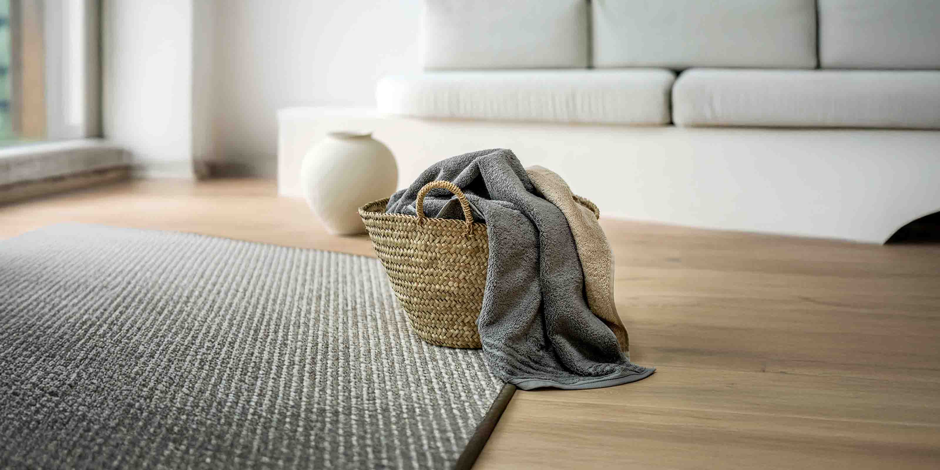 Wohnzimmer mit Korb voller gefalteter Decken oder Handtücher auf Holzboden, grauer Teppich, weißes Sofa mit Kissen und weiße Vase. Tageslicht von links.