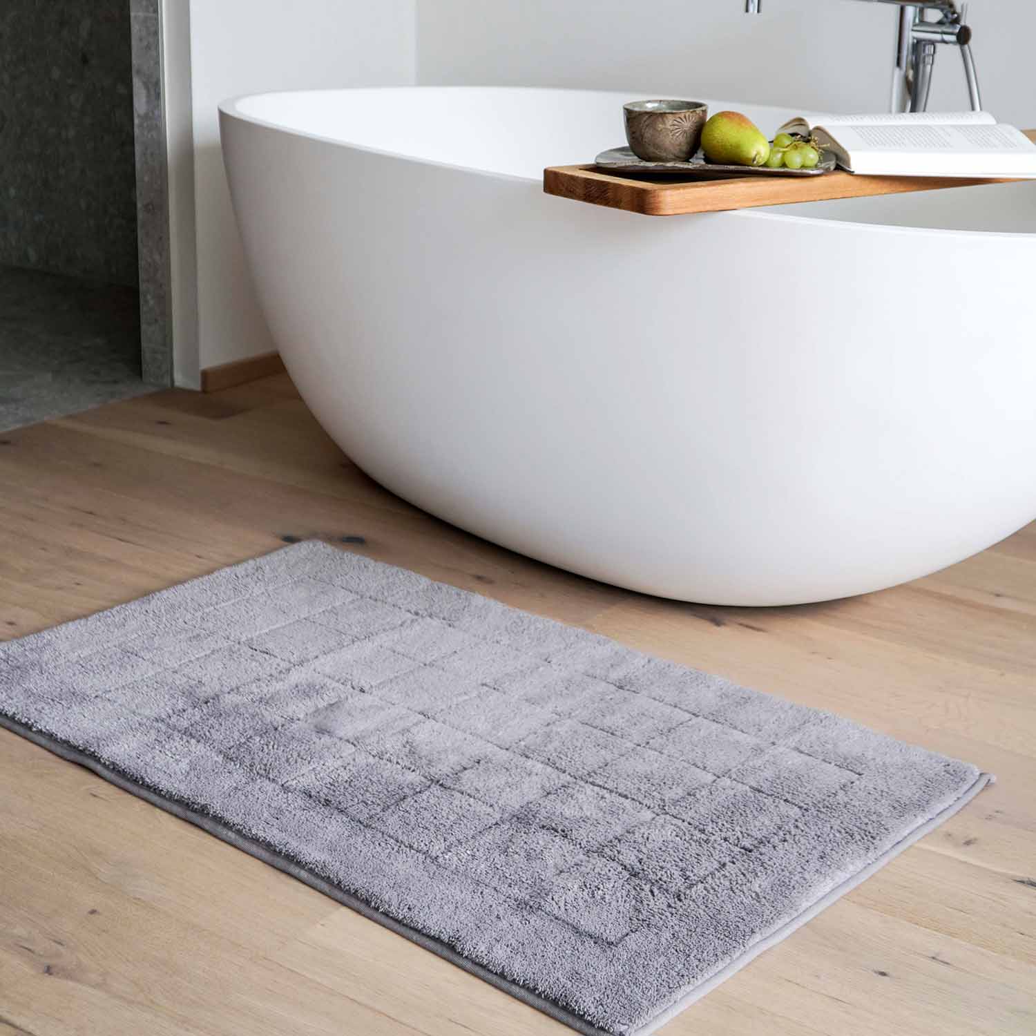Modernes Badezimmer mit freistehender Badewanne, Holzablage mit Obst, Tasse und Buch, davor graue Badematte auf Holzboden.