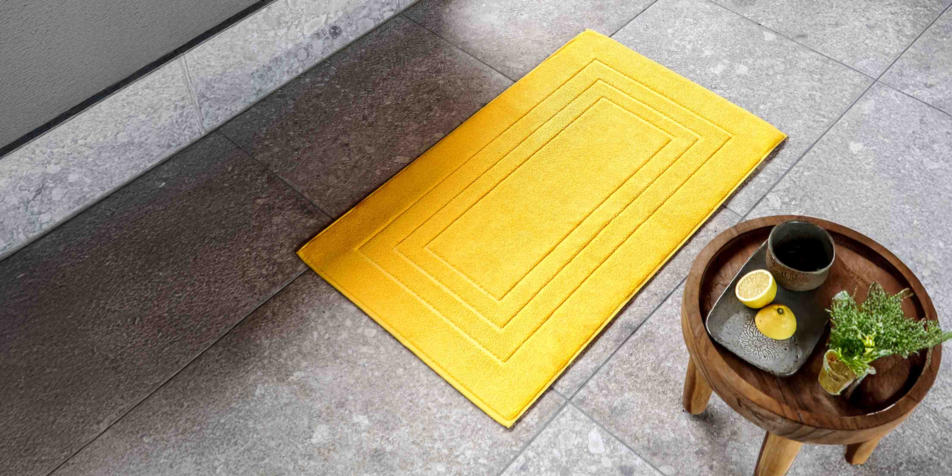 Gelber Teppich auf grauem Fliesenboden neben rundem Holztisch mit Getränk und Zitrone.