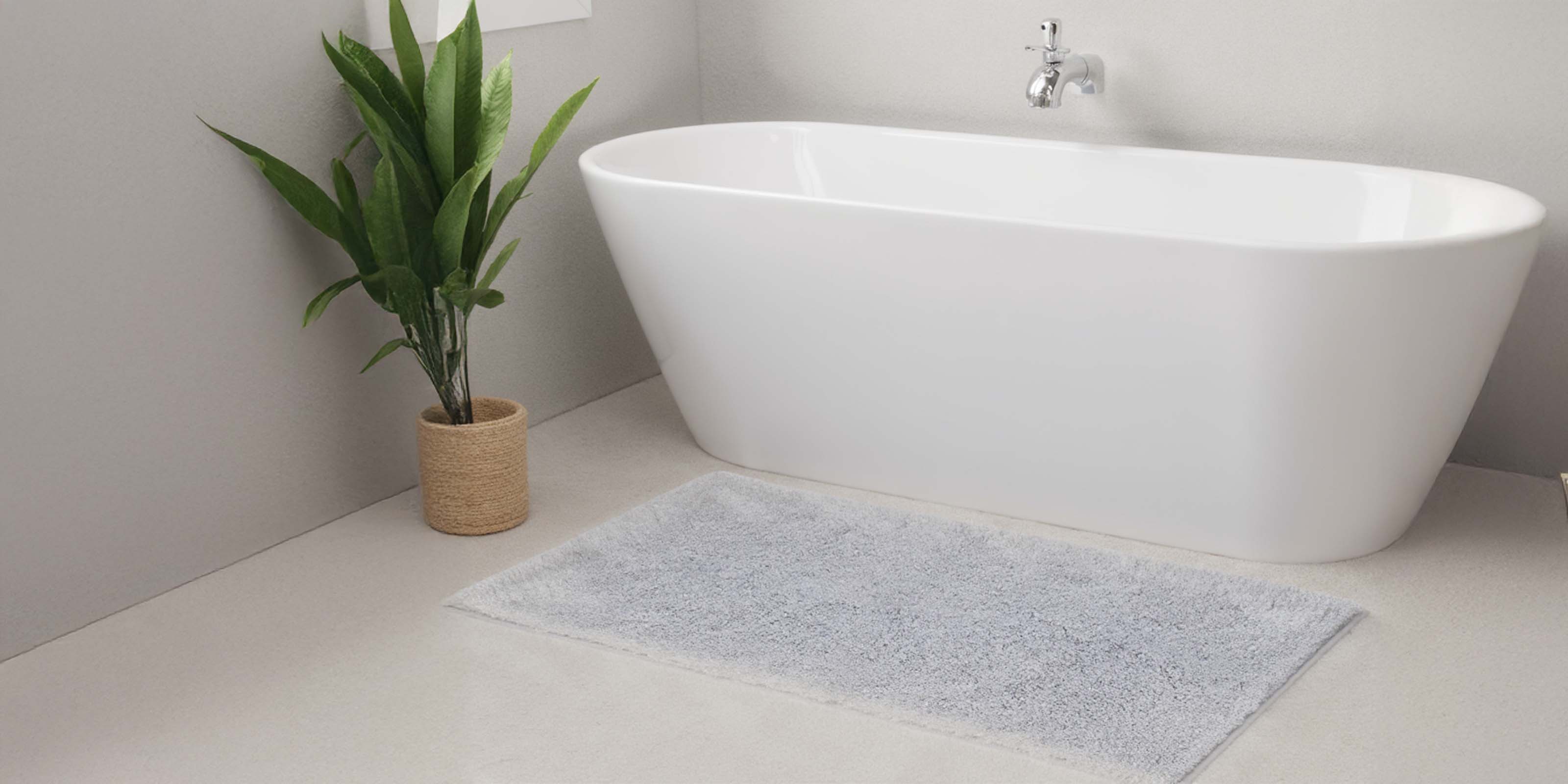Modernes Badezimmer mit freistehender weißer Badewanne, Chromarmatur, grünem Blumentopf im Korb und hellgrauer Badematte auf dem Boden.