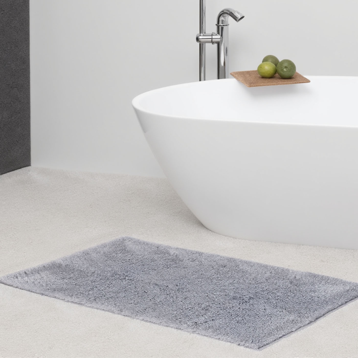 Modernes Badezimmer mit freistehender weißer Badewanne und Wandarmatur. Auf dem Wannenrand liegt ein Holztablett mit drei Limetten. Davor eine graue Badematte auf hellem Boden.