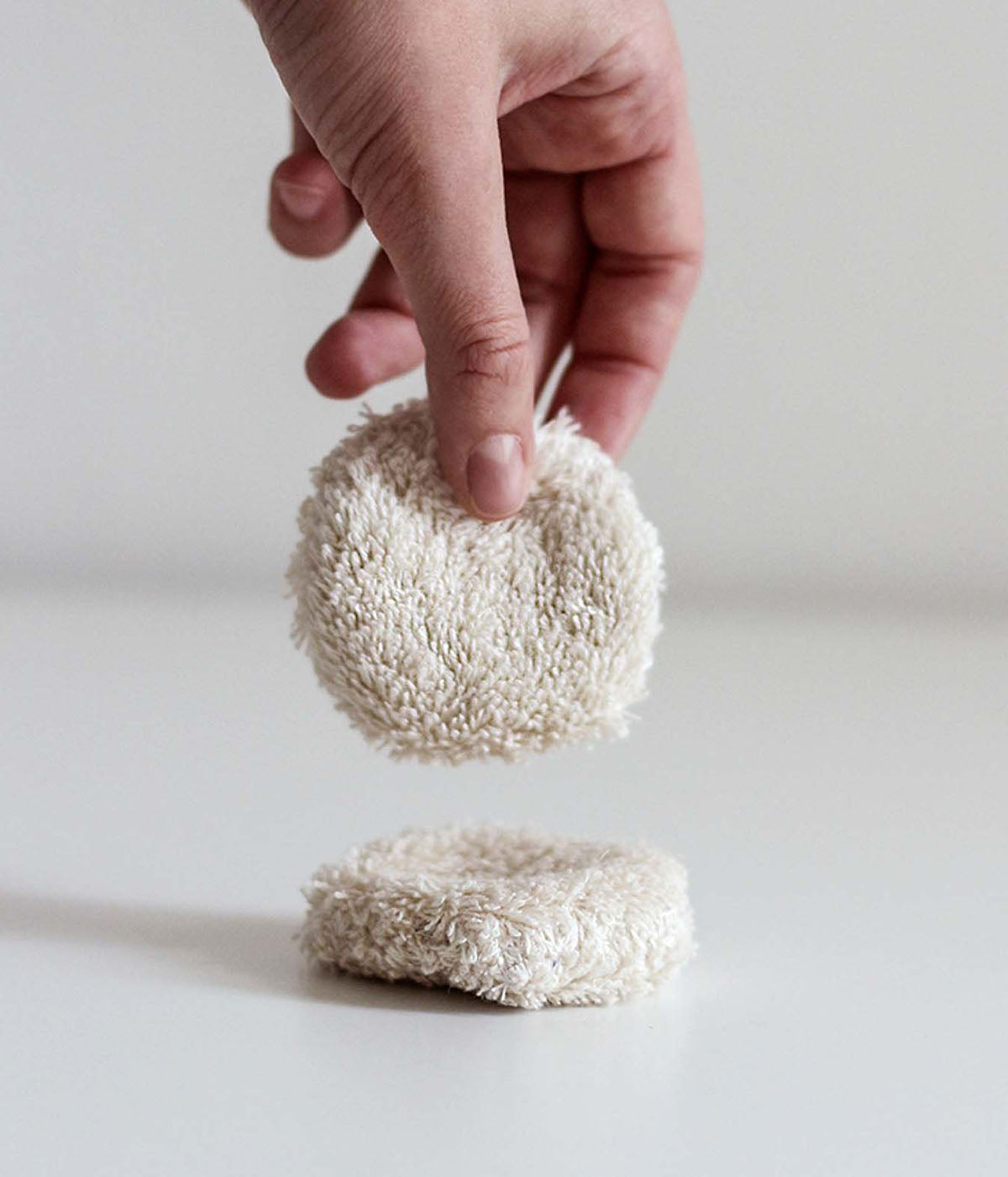 Eine Hand hält ein rundes, beigefarbenes, wiederverwendbares Abschminkpad. Dasselbe liegt darunter auf einer hellen Fläche.