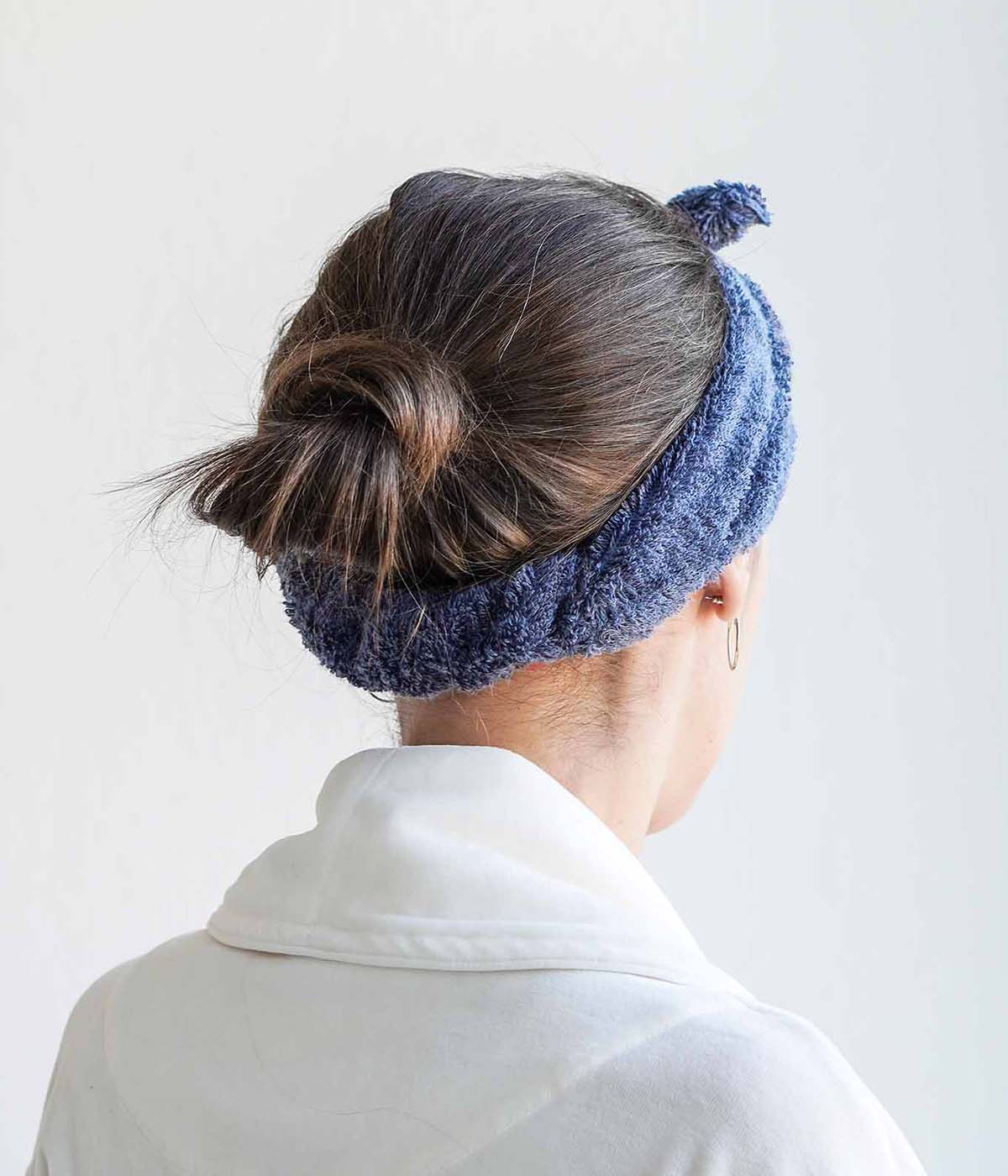 Rückansicht einer Person mit weißem Bademantel und blauem Haarband. Die Haare sind zu einem Dutt gebunden.
