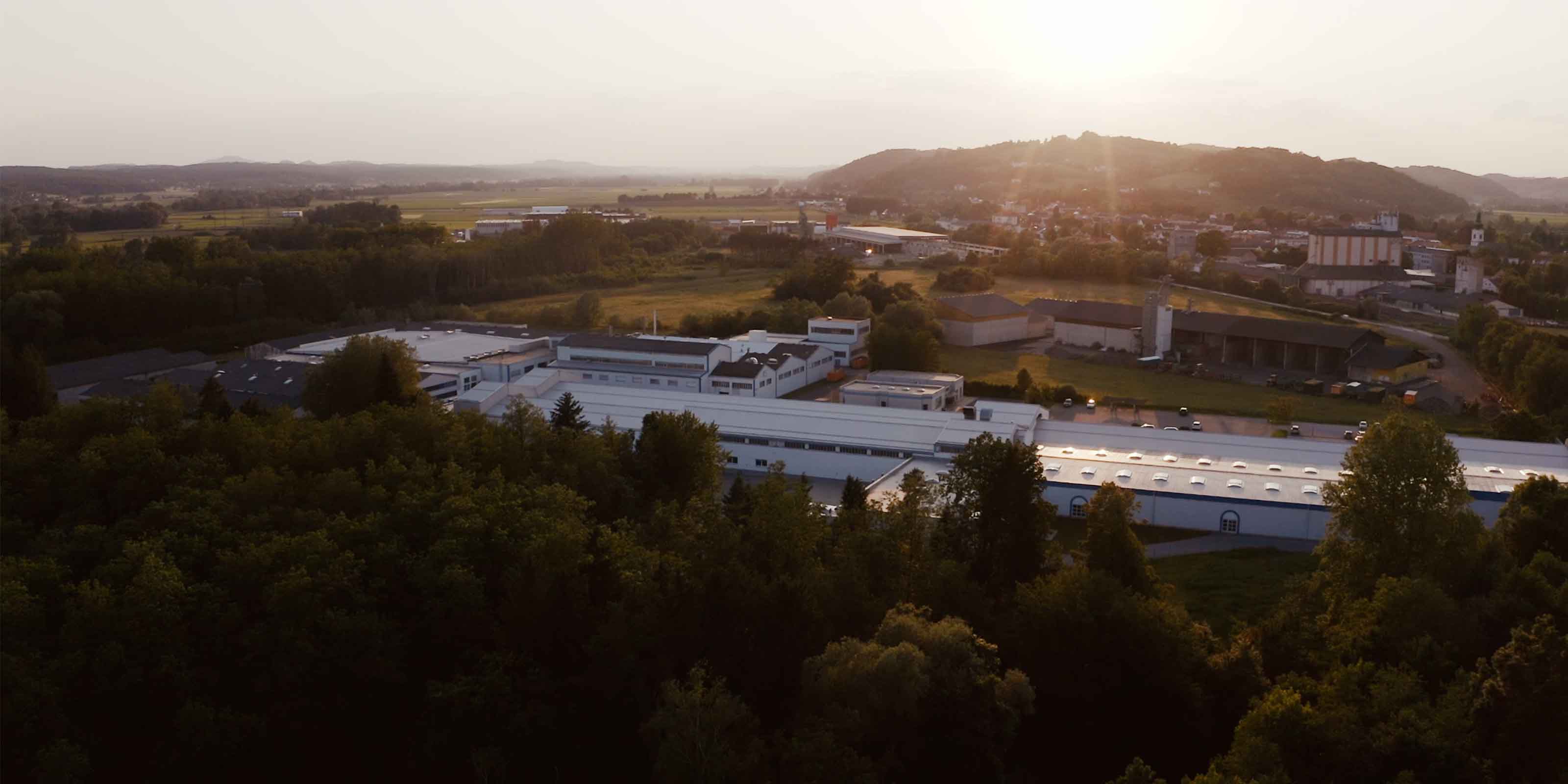 Luftaufnahme eines Industriegebäudes im Grünen bei Sonnenuntergang; im Hintergrund eine Stadt mit Hügeln.