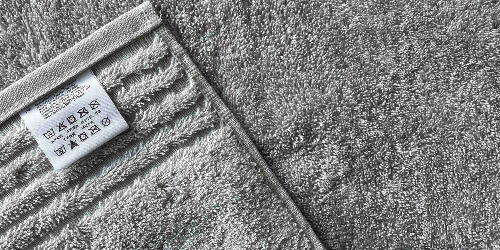 Detailaufnahme eines Pflegeetiketts auf einem strukturierten grauen Handtuch mit Wasch- und Pflegesymbolen.