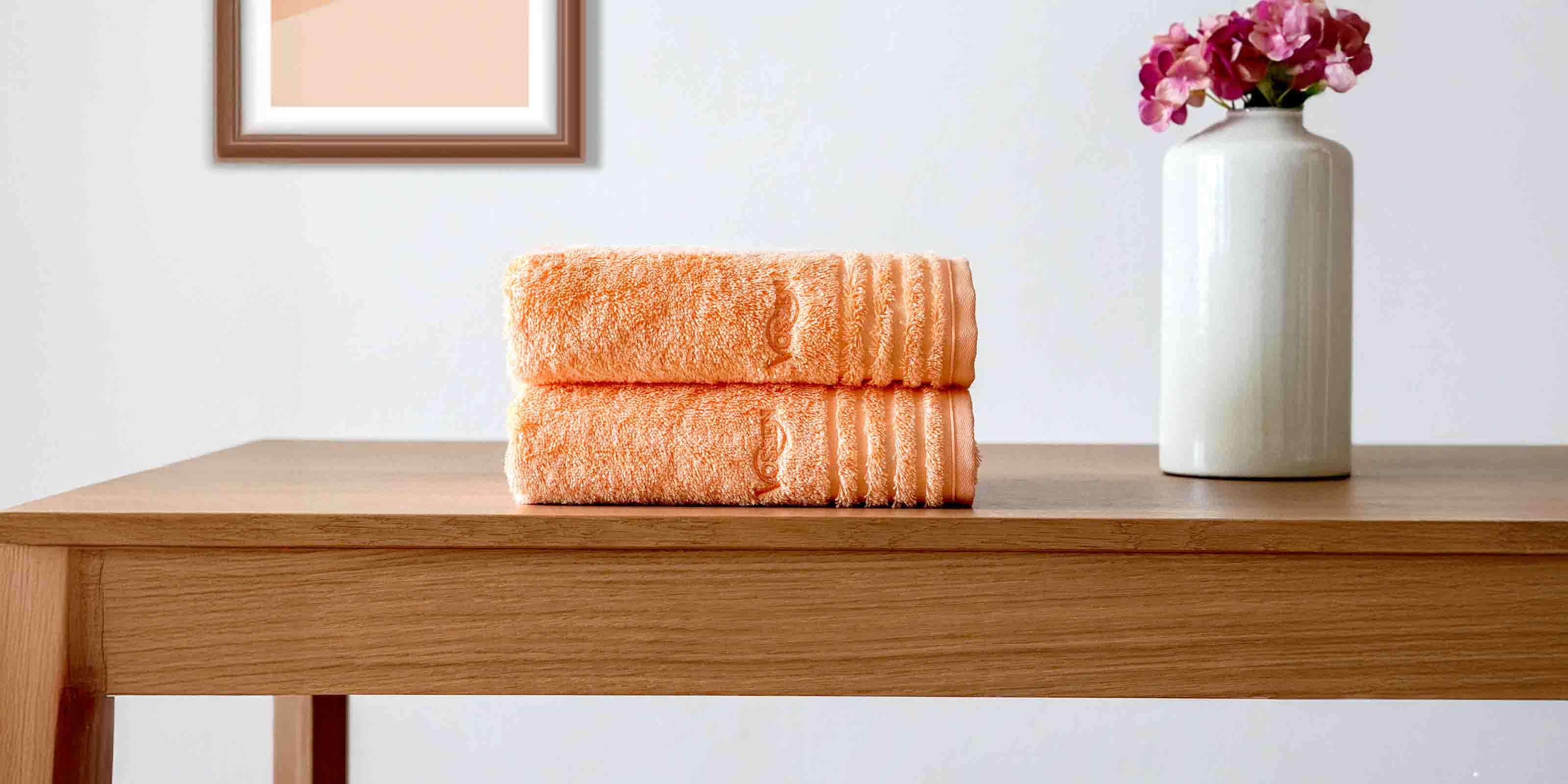 Zwei orangefarbene Handtücher übereinander zusammengelegt liegend auf einem Holztisch, daneben eine weiße Vase mit pinken Blumen.