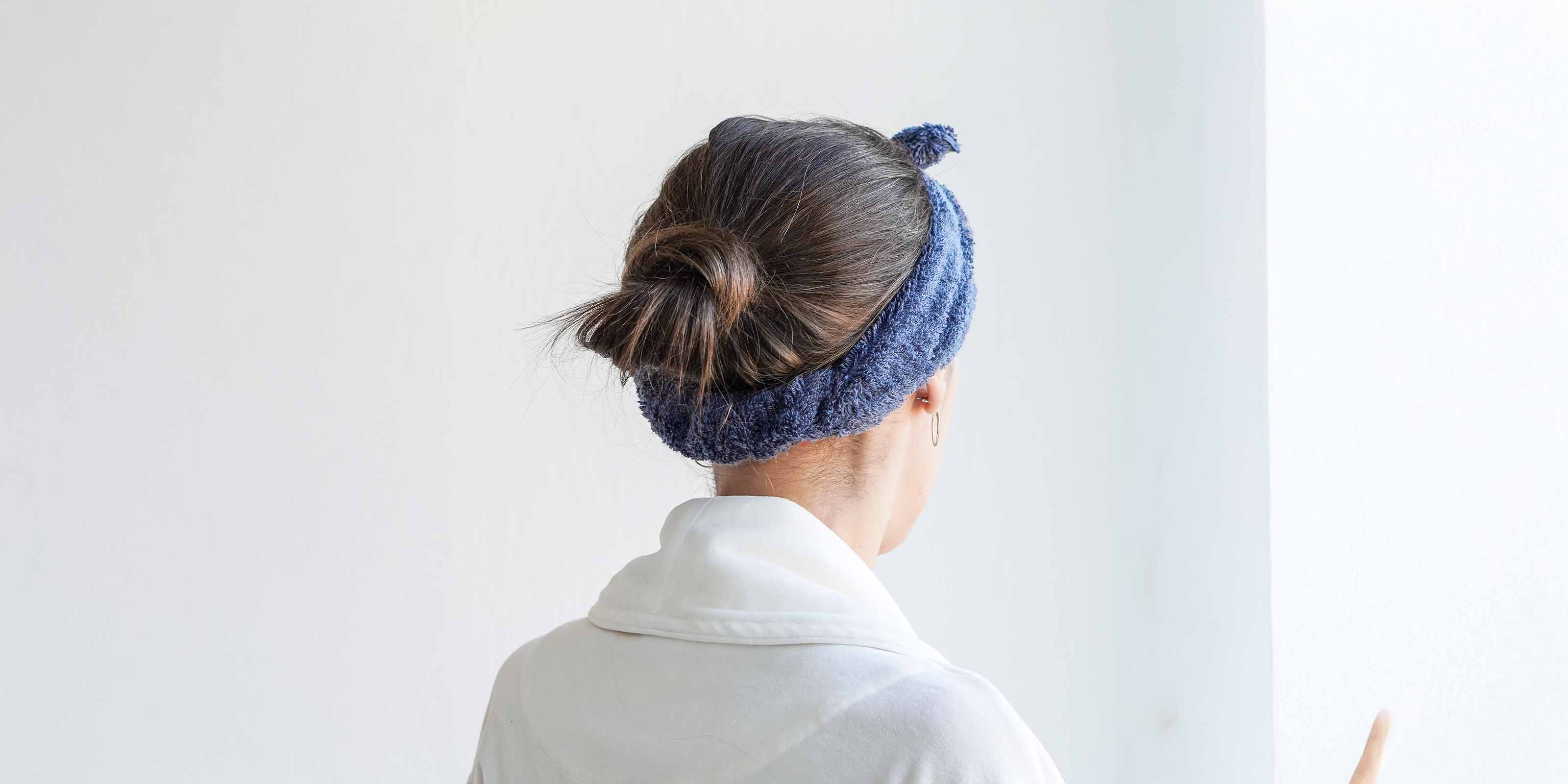 Rückansicht einer Person mit weißem Bademantel und blauem Haarband. Die Haare sind zu einem Dutt gebunden.