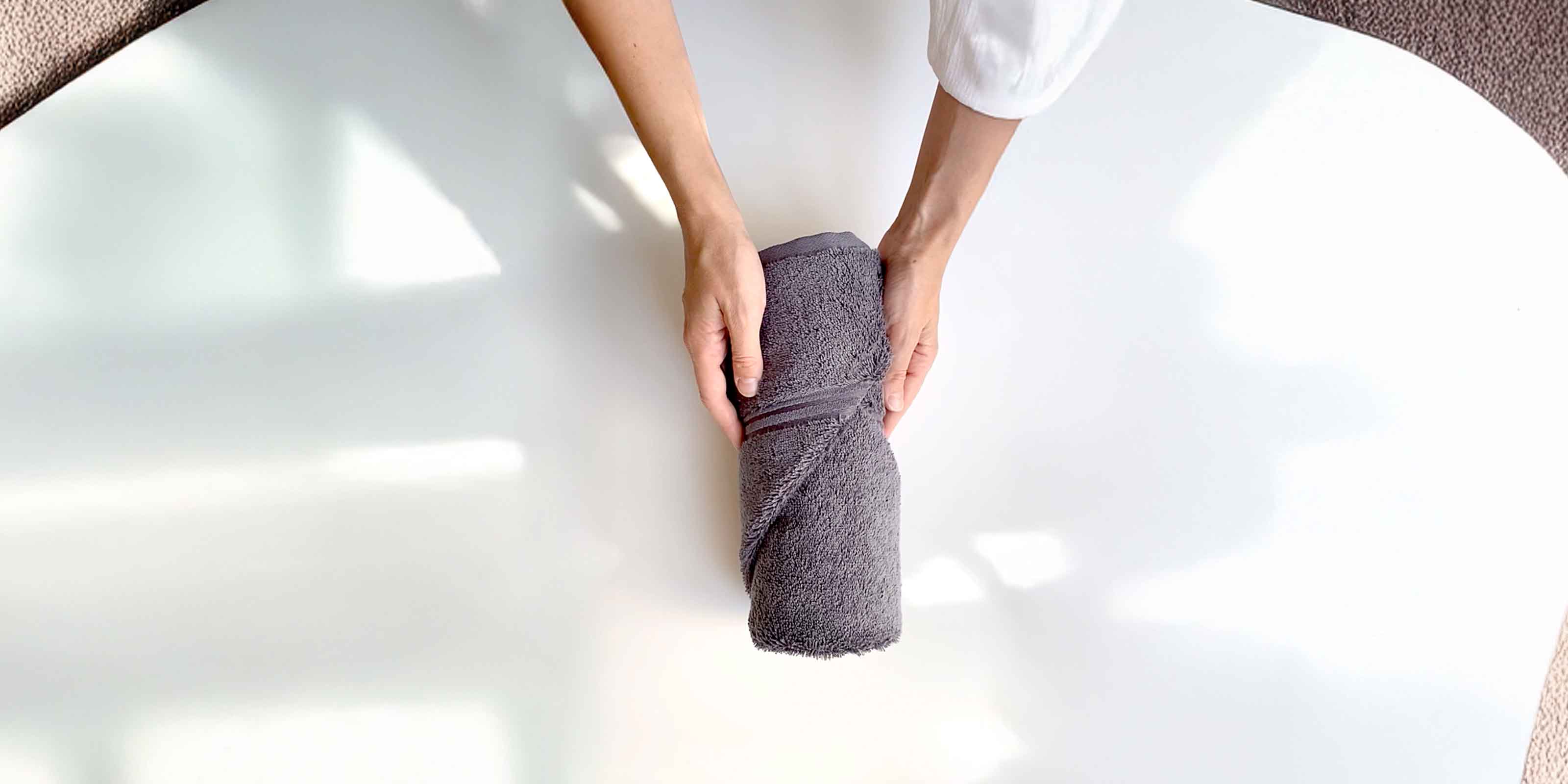 Zwei Hände halten ein ordentlich zusammengerolltes graues Handtuch vor hellem Hintergrund.