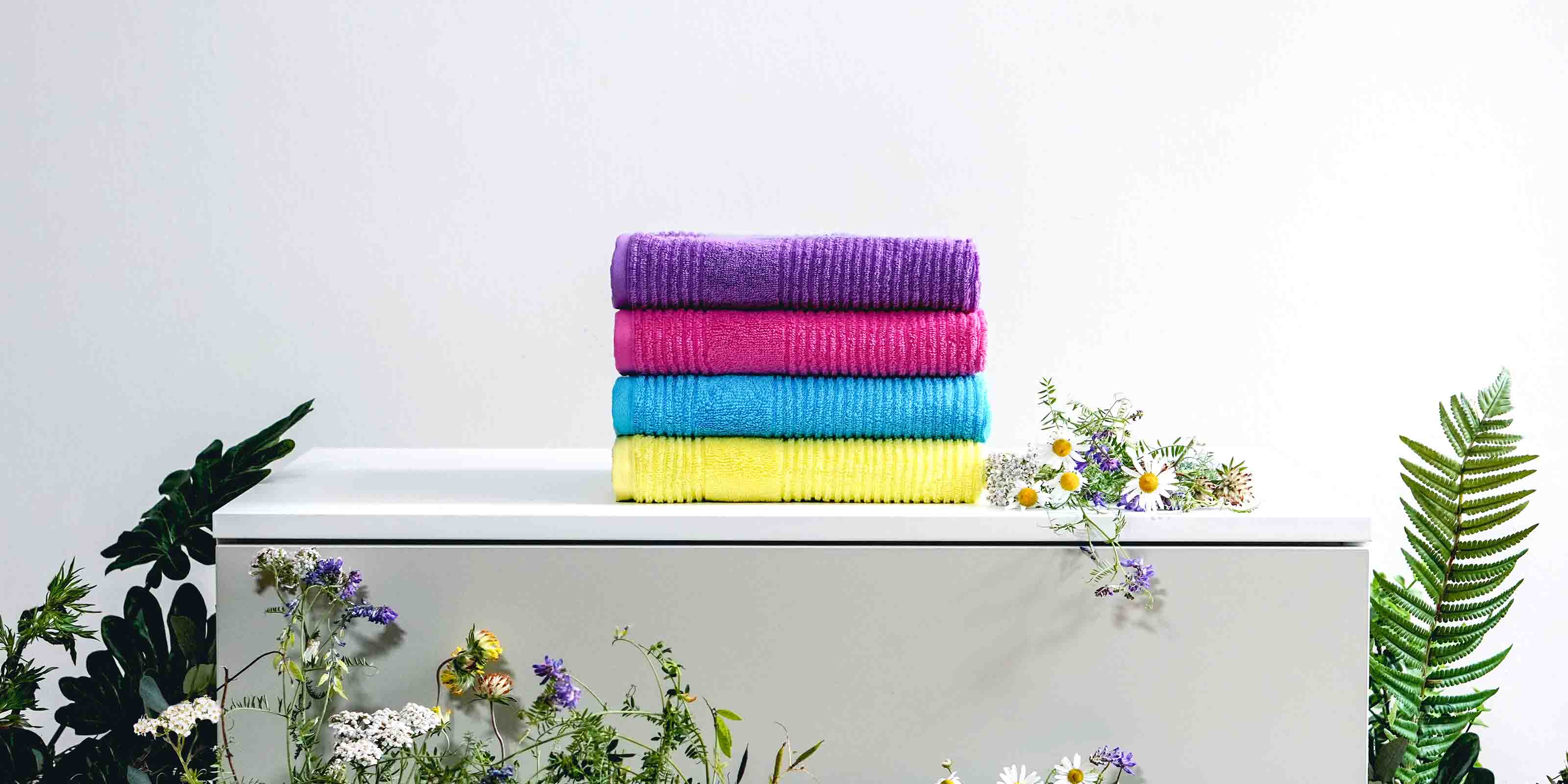 Vier farbenfrohe, gestapelte Handtücher in Lila, Pink, Blau und Gelb vor hellgrauem Hintergrund. Die Szene ist mit frischen Wiesenblumen wie Gänseblümchen, Klee und anderen Wildblumen dekoriert.