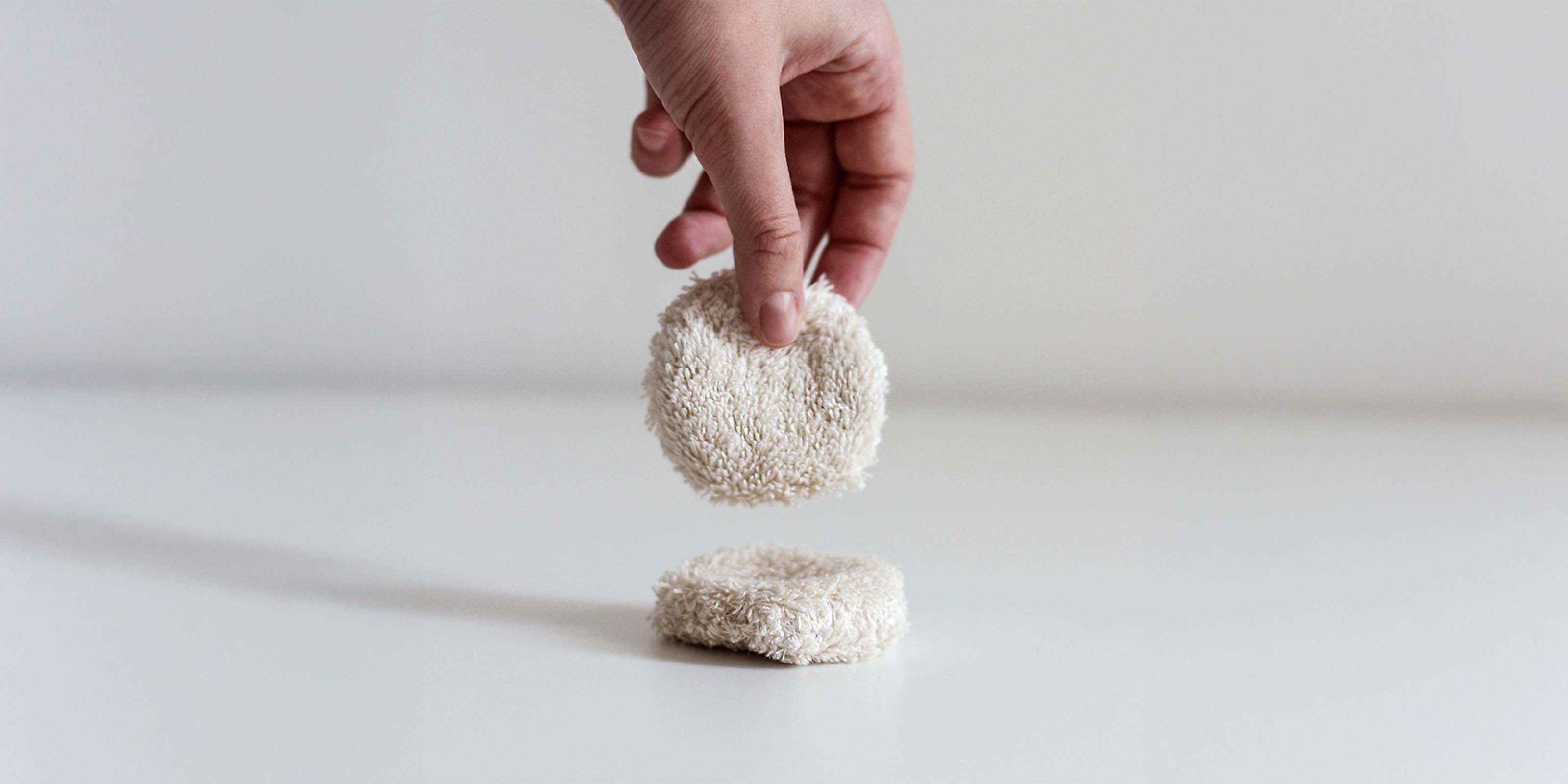 Eine Hand hält ein rundes, beigefarbenes, wiederverwendbares Abschminkpad. Dasselbe liegt darunter auf einer hellen Fläche.