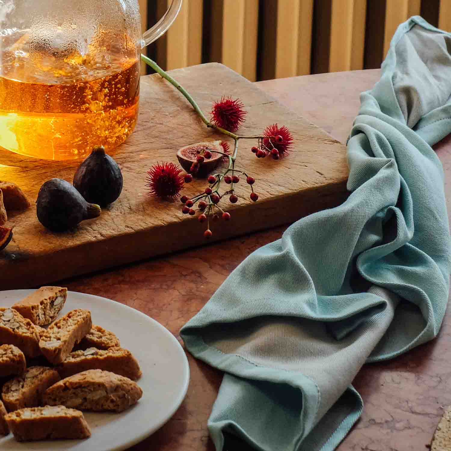 Stilvolles Stillleben mit Glaskanne Tee, Feigen, Gebäck und dekorativen Beeren auf Holzbrett – daneben liegt ein mintgrünes Küchentuch auf dem Tisch