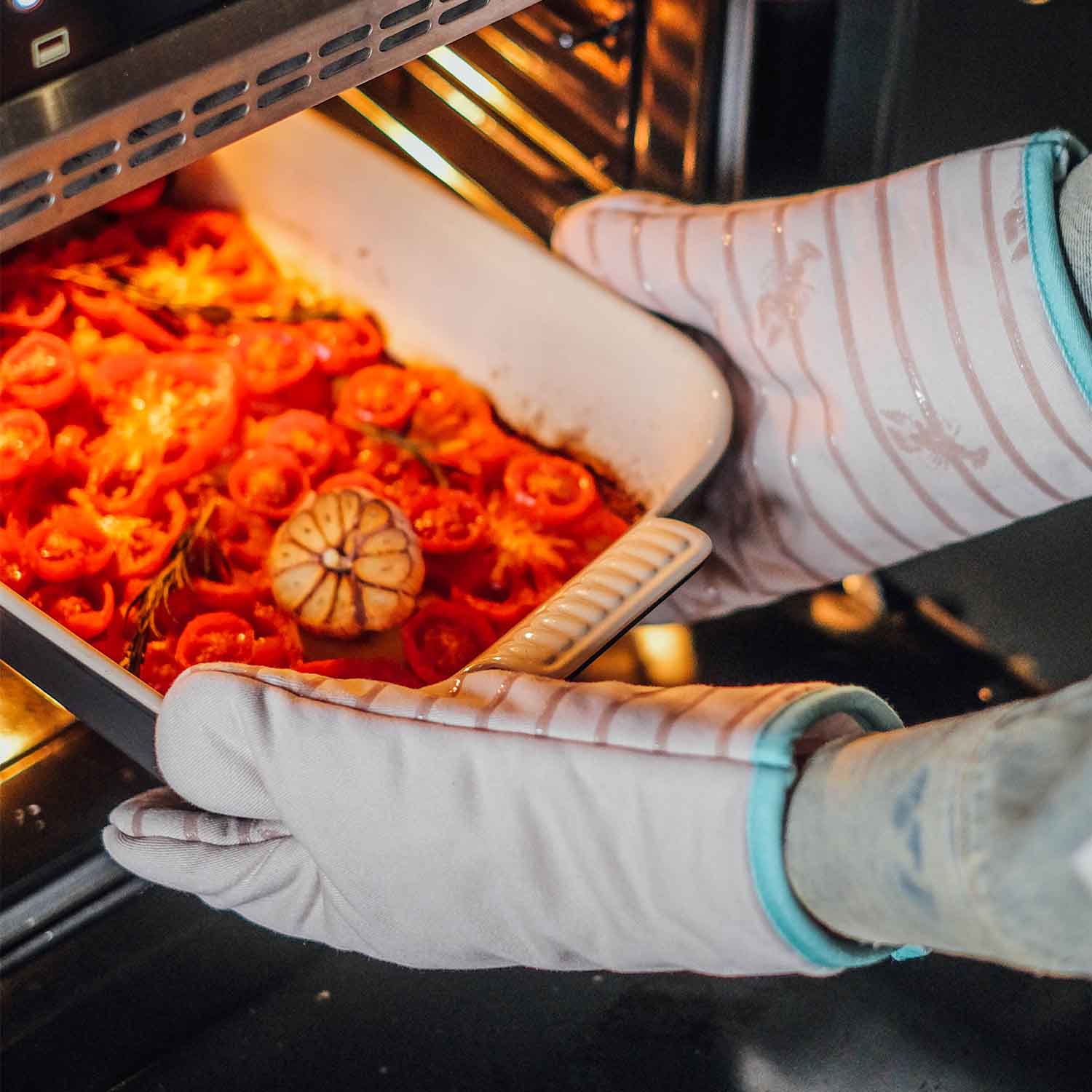 Person mit gestreiften Ofenhandschuhen holt Auflaufform mit gerösteten Tomaten aus dem Backofen – hitzebeständige Küchenhandschuhe für sicheres Backen und Kochen