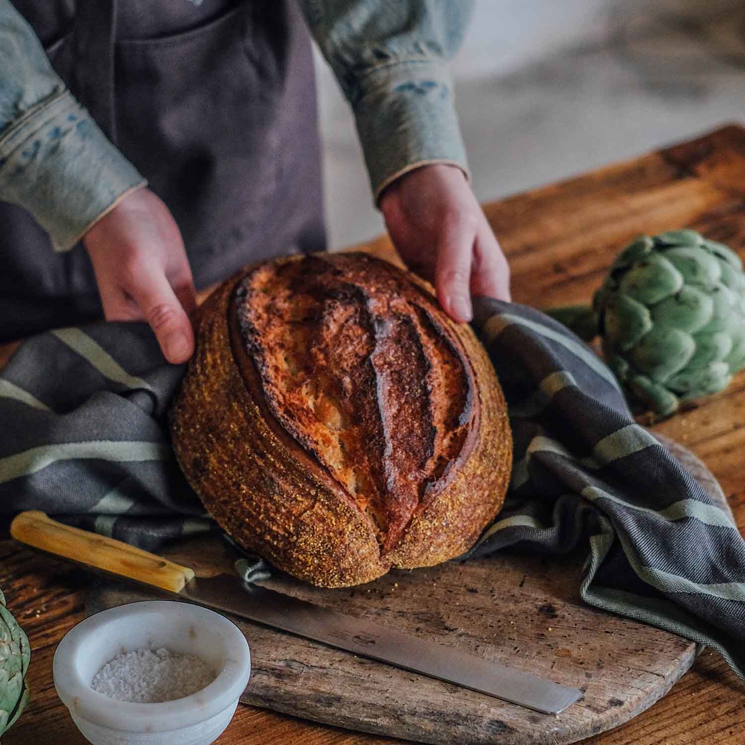 Frisch gebackenes Bauernbrot auf rustikalem Holzbrett, gehalten von Händen mit Küchentuch – daneben Salzschale, Brotmesser und Artischocke auf Holztisch