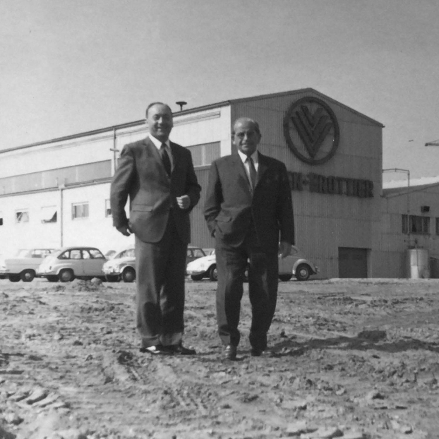 Zwei Männer in Anzügen stehen vor einem großen Industriegebäude mit ‚V‘-Schriftzug, daneben mehrere geparkte Autos.