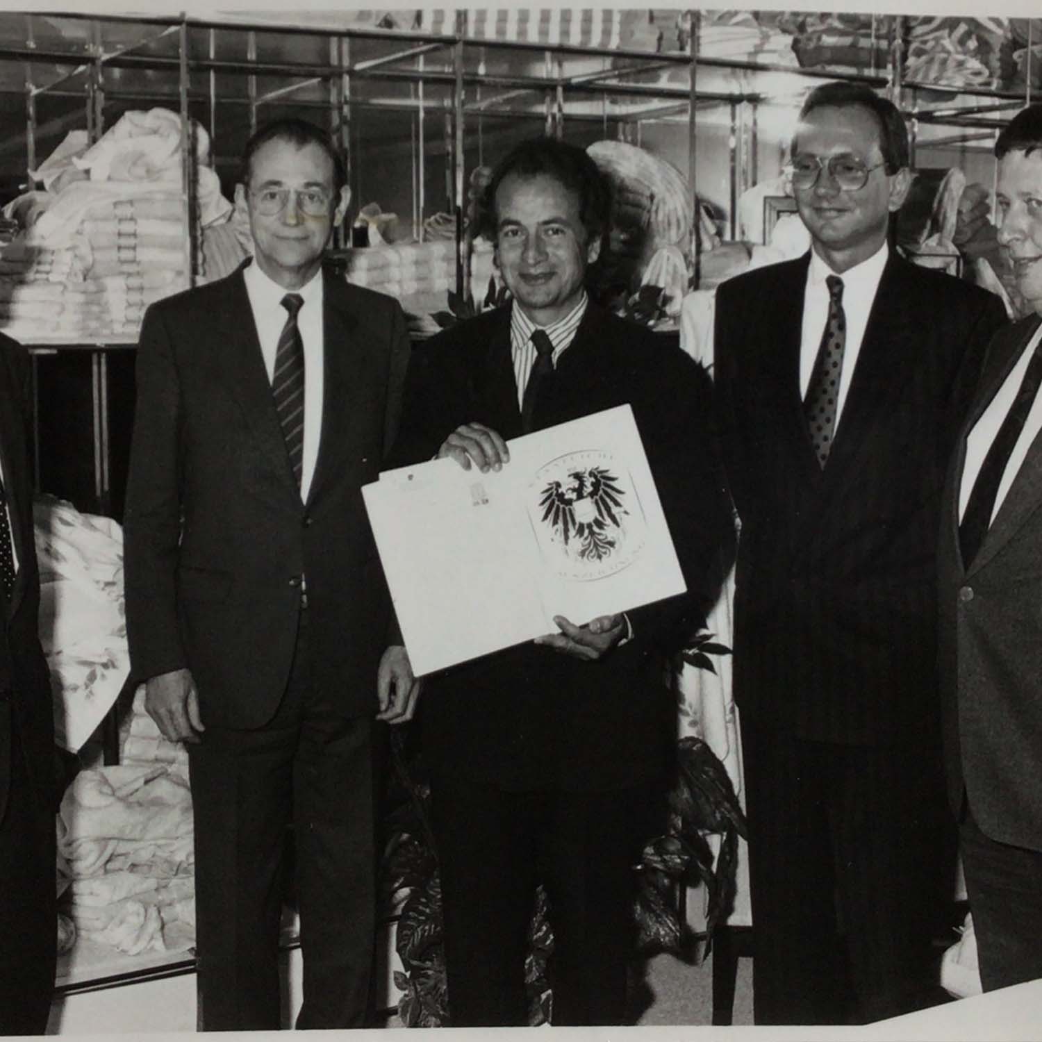 Gruppe von Männern in Anzügen, einer hält ein geöffnetes Dokument mit Adler-Wappen, Hintergrund mit Regalen.