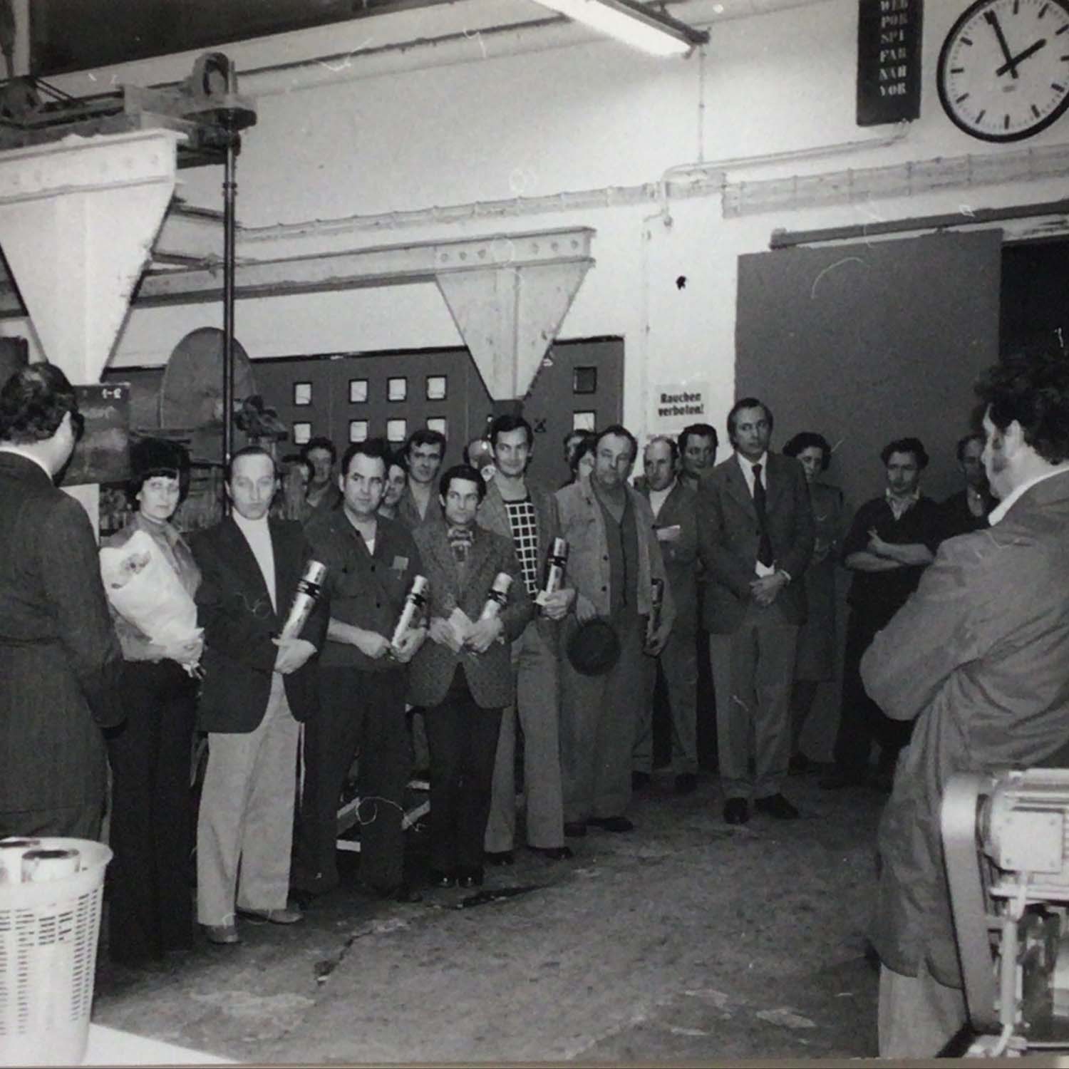 Gruppe von Personen in formeller Kleidung in Fabrikumgebung, einige halten Zylinderobjekte, Maschinen im Hintergrund.