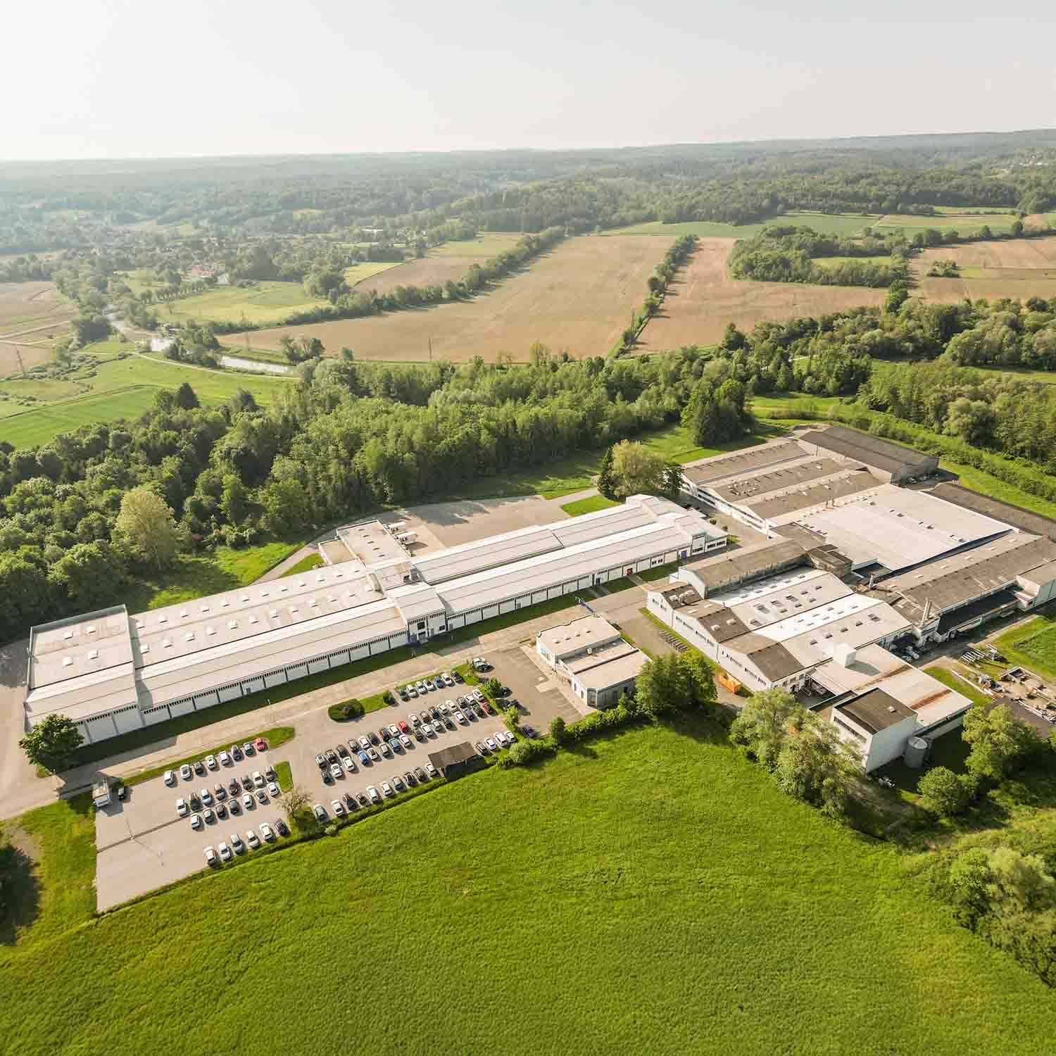 Ein Industriegebäude mit Parkplätzen von oben abgelichtet inmitten von Feldern und grünen Wäldern.