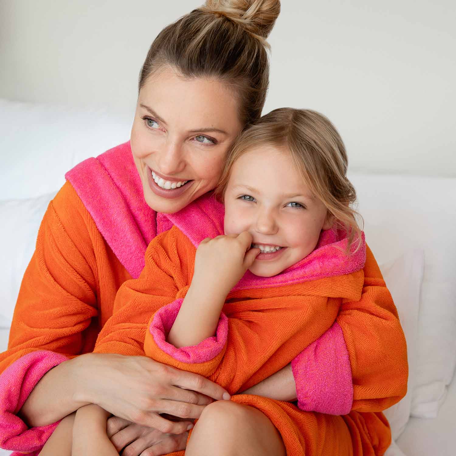 Frau umarmt lächelnd Kind in orange-pinken Bademänteln.