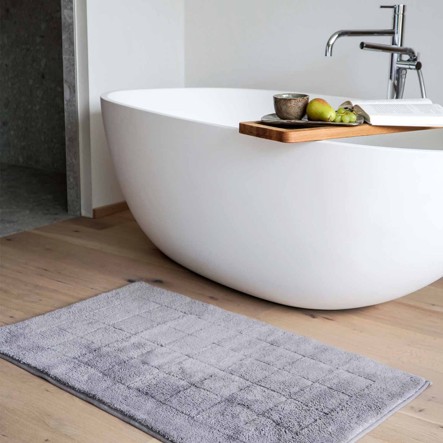 Freistehende Badewanne auf Holzboden, davor liegt ein grauer Frottierteppich.