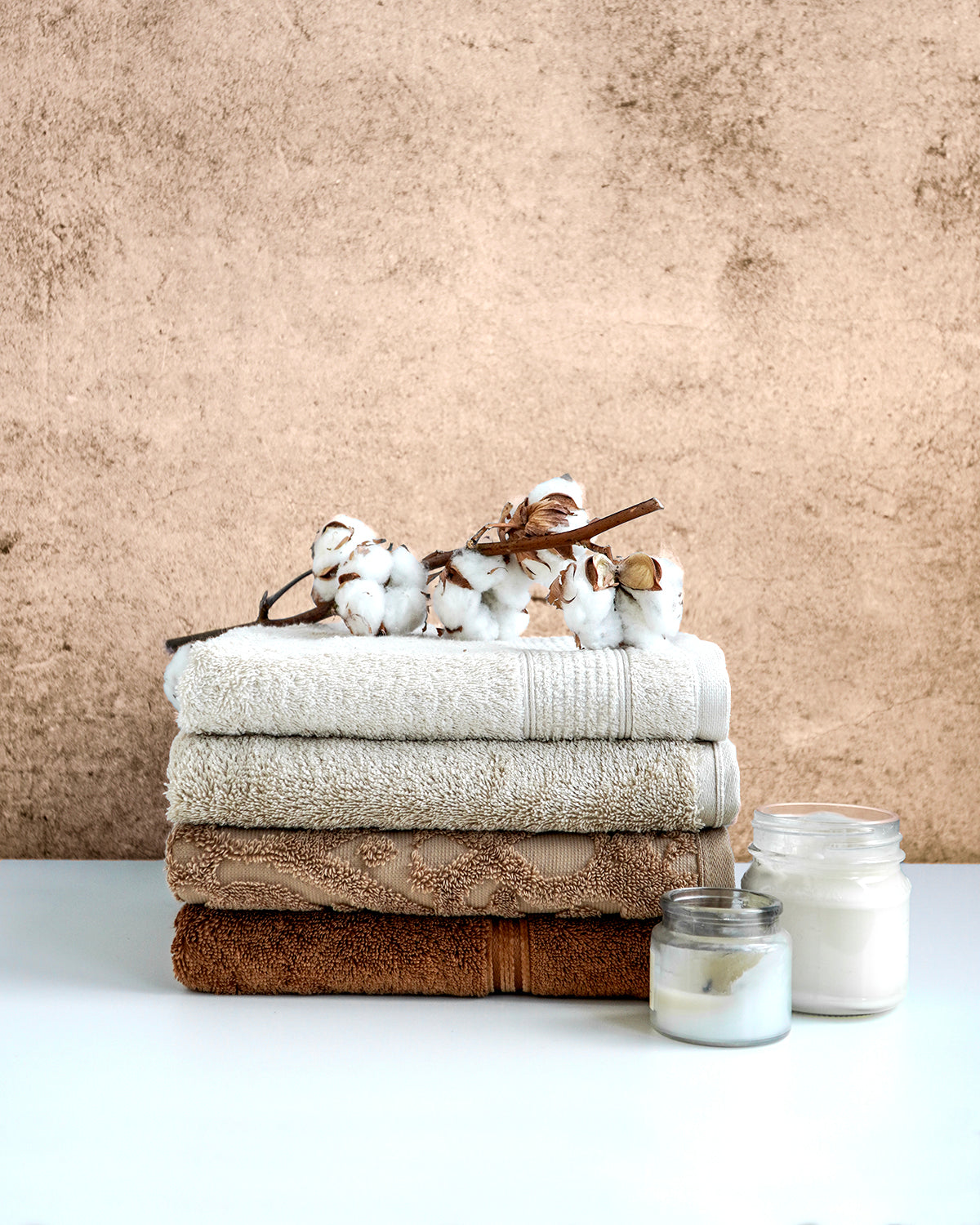 Gefaltete Handtücher in Naturtönen mit Baumwollzweigen und Duftkerzen auf weißer Oberfläche vor beiger Wand für ein gemütliches Wellnessambiente.
