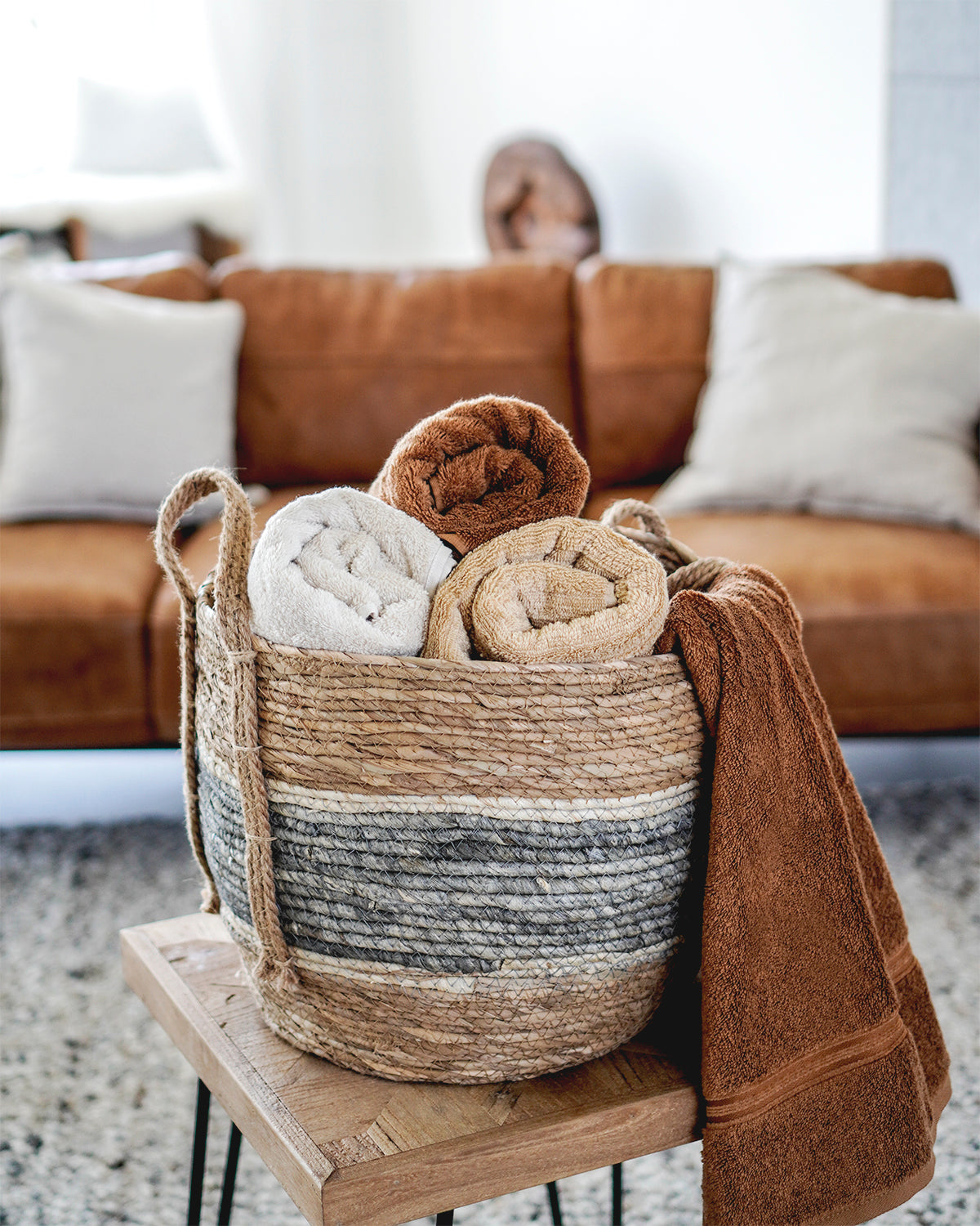 Geflochtener Korb mit zusammengerollten Handtüchern in warmen Naturtönen steht auf einem Holztisch vor einem braunen Sofa im gemütlichen Wohnzimmer.