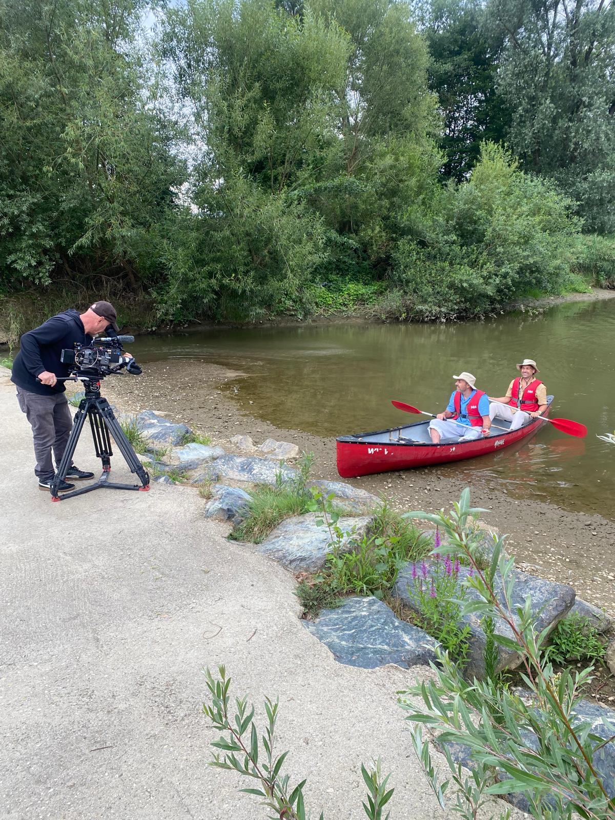 Zwei Männer in einem roten Kanu am Flussufer, während ein Kameramann die Szene mit einer professionellen Filmkamera aufnimmt.