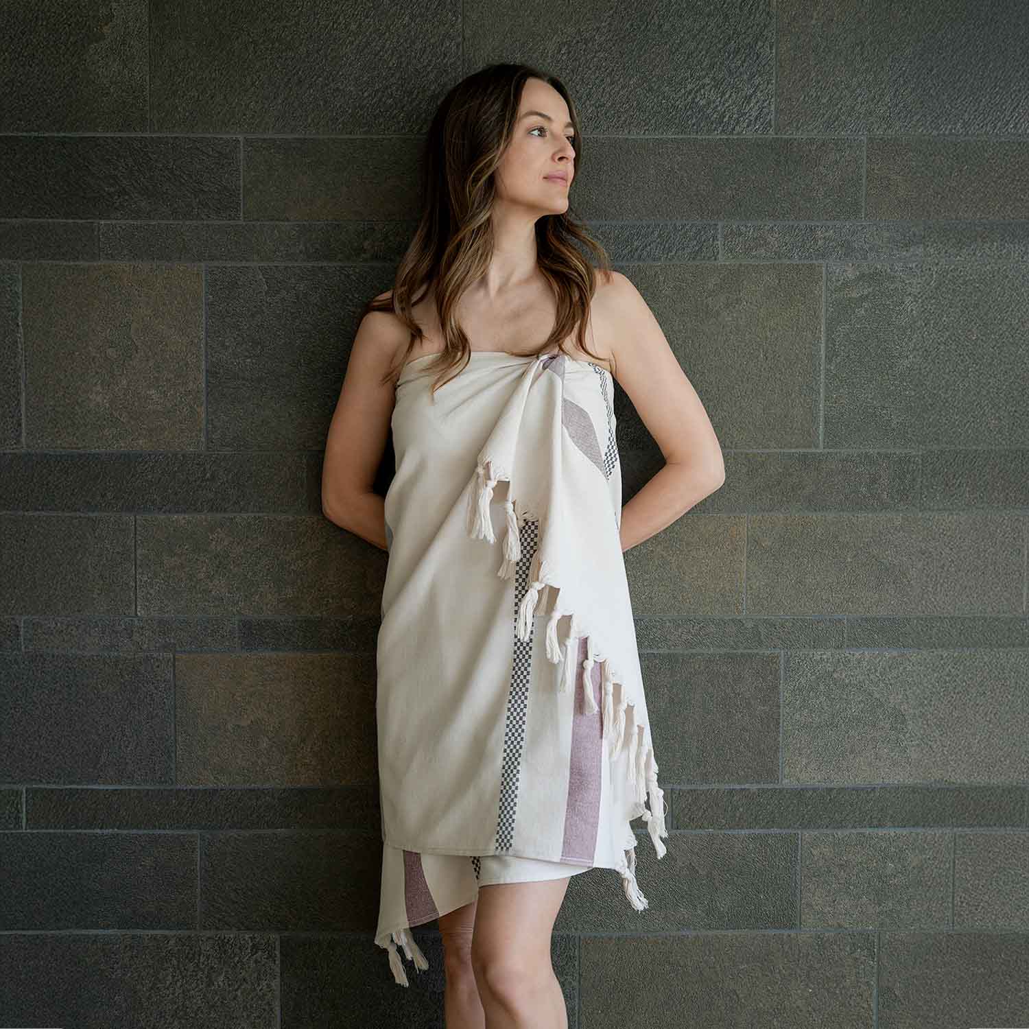 Frau steht entspannt vor einer Steinwand und trägt ein helles Hamam-Tuch mit dezenten Streifen und Fransen – Sinnbild für Natürlichkeit und Wellness.