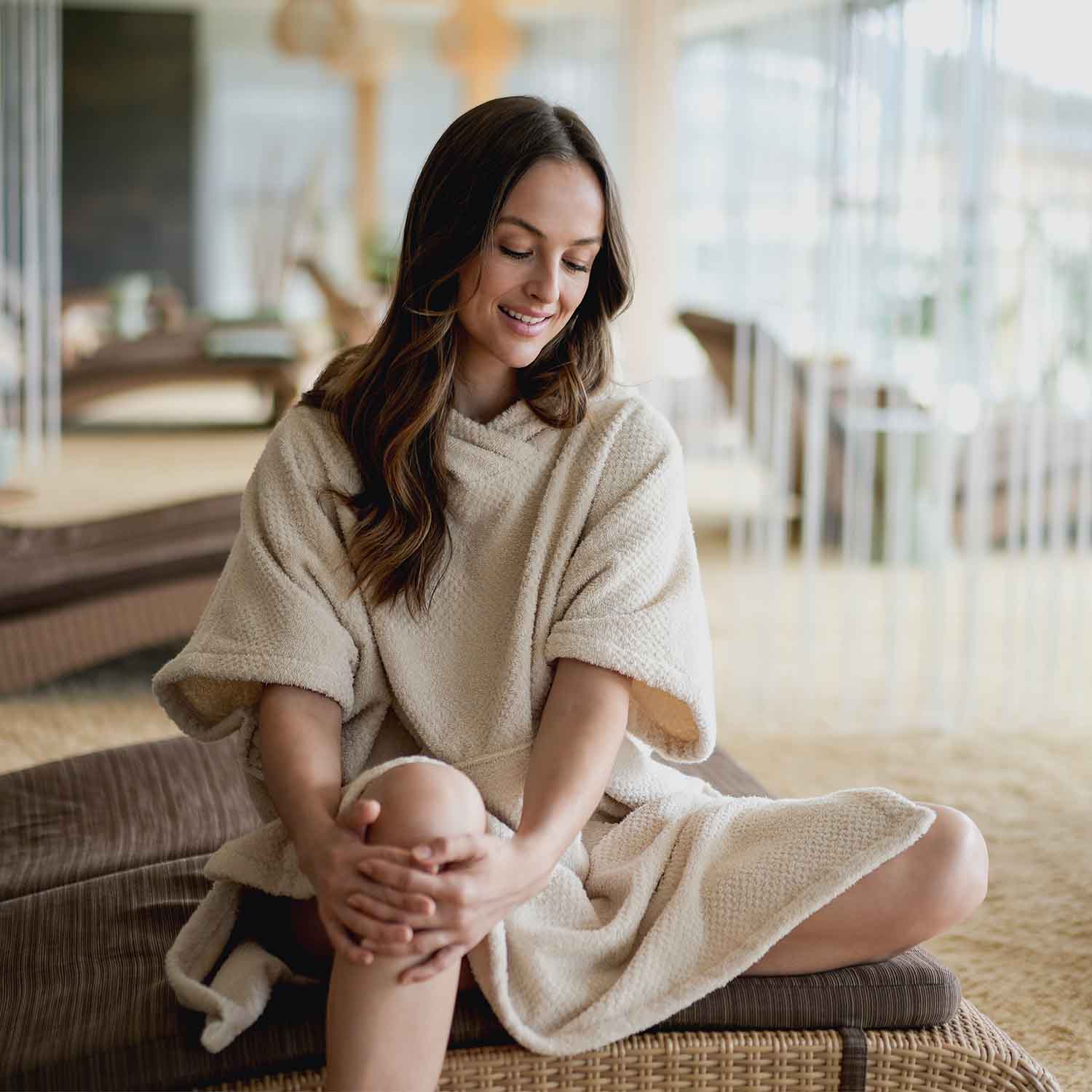 Frau entspannt in einem cremefarbenen Kapuzenponcho aus Frottee im Wellnessbereich und genießt eine ruhige Auszeit.