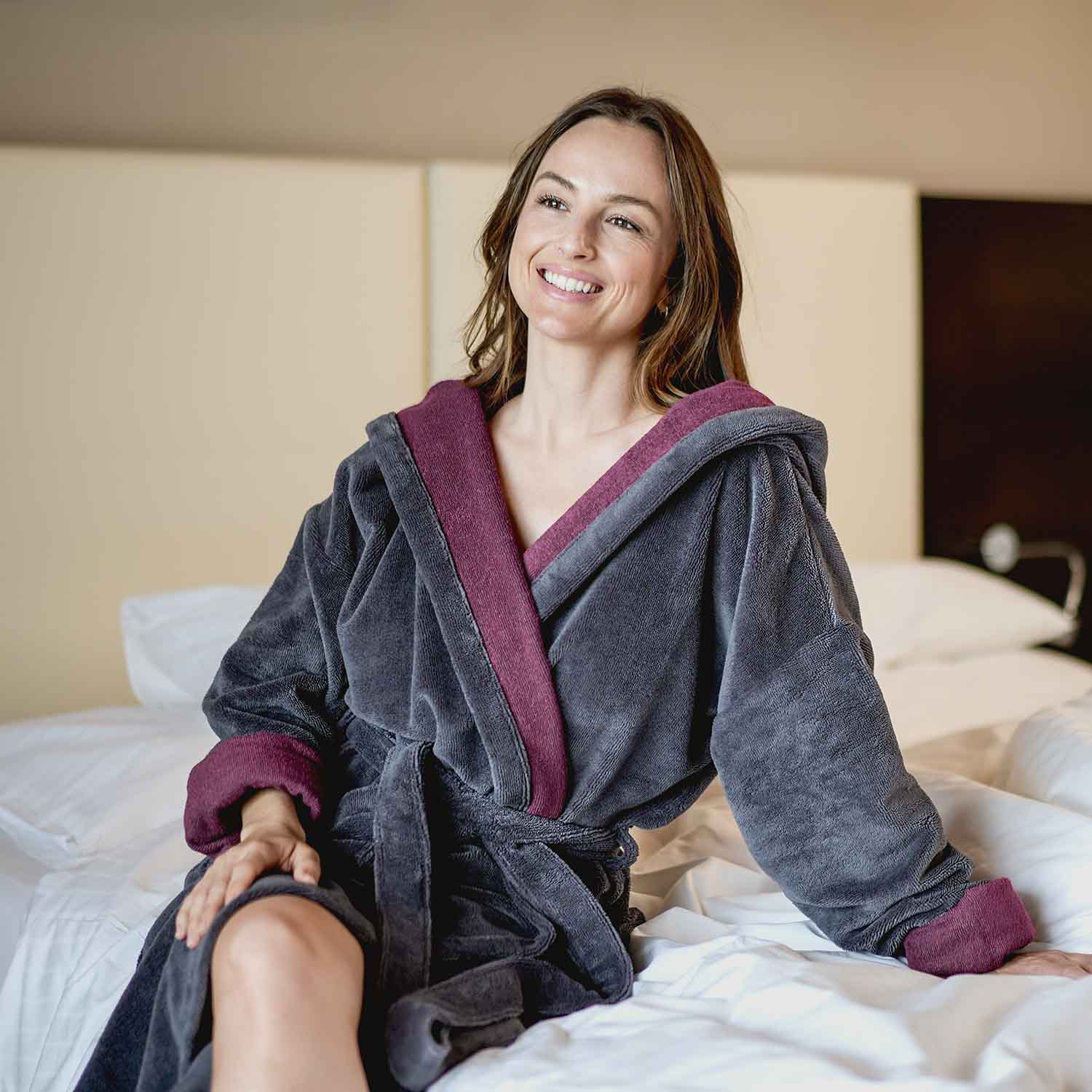 Frau sitzt lächelnd auf einem Bett und trägt einen grauen Bademantel mit weinrotem Innenfutter – Symbol für Komfort und Wohlfühlen im Alltag.