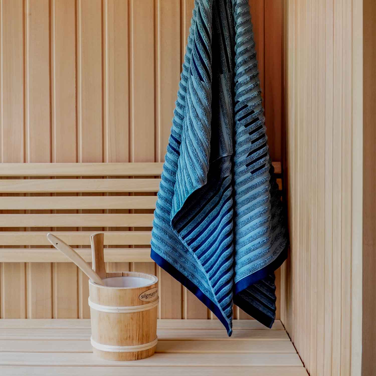 Blaues Handtuch mit Streifenstruktur hängt in einer hellen Holzsauna neben einem Aufgusseimer aus Holz – Sinnbild für Wellness und Entspannung.