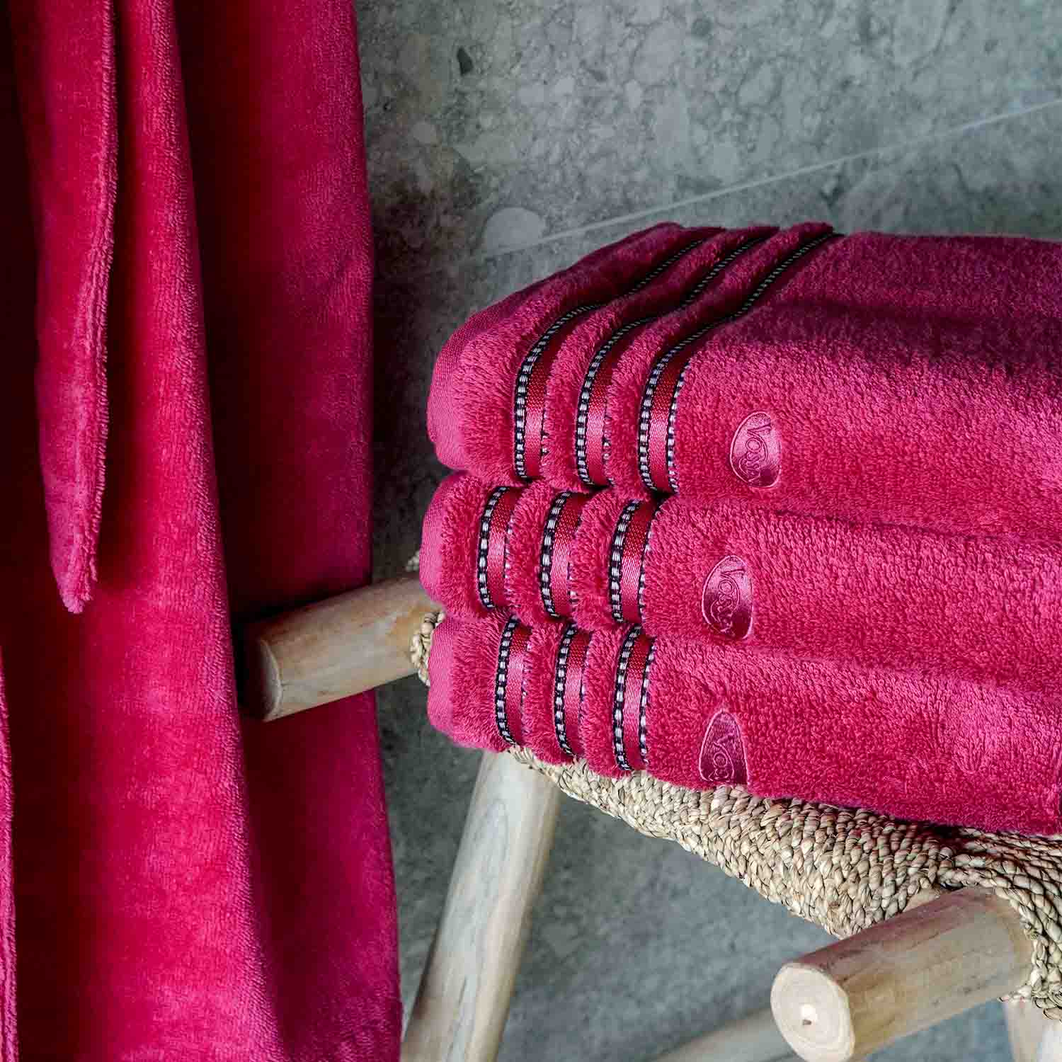 Vier pink gefaltete Handtücher liegen auf einem Holzregal. Links hängt ein pinker Bademantel. Die Handtücher haben Ziernähte, im Hintergrund ist eine Stein- oder Fliesenwand.