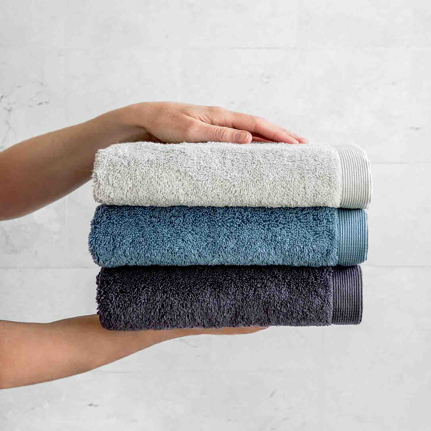 Drei gefaltete Handtücher in Grau-, Blau- und Grautönen vor einer hellen Wand, gehalten von zwei Händen, oben und unten.