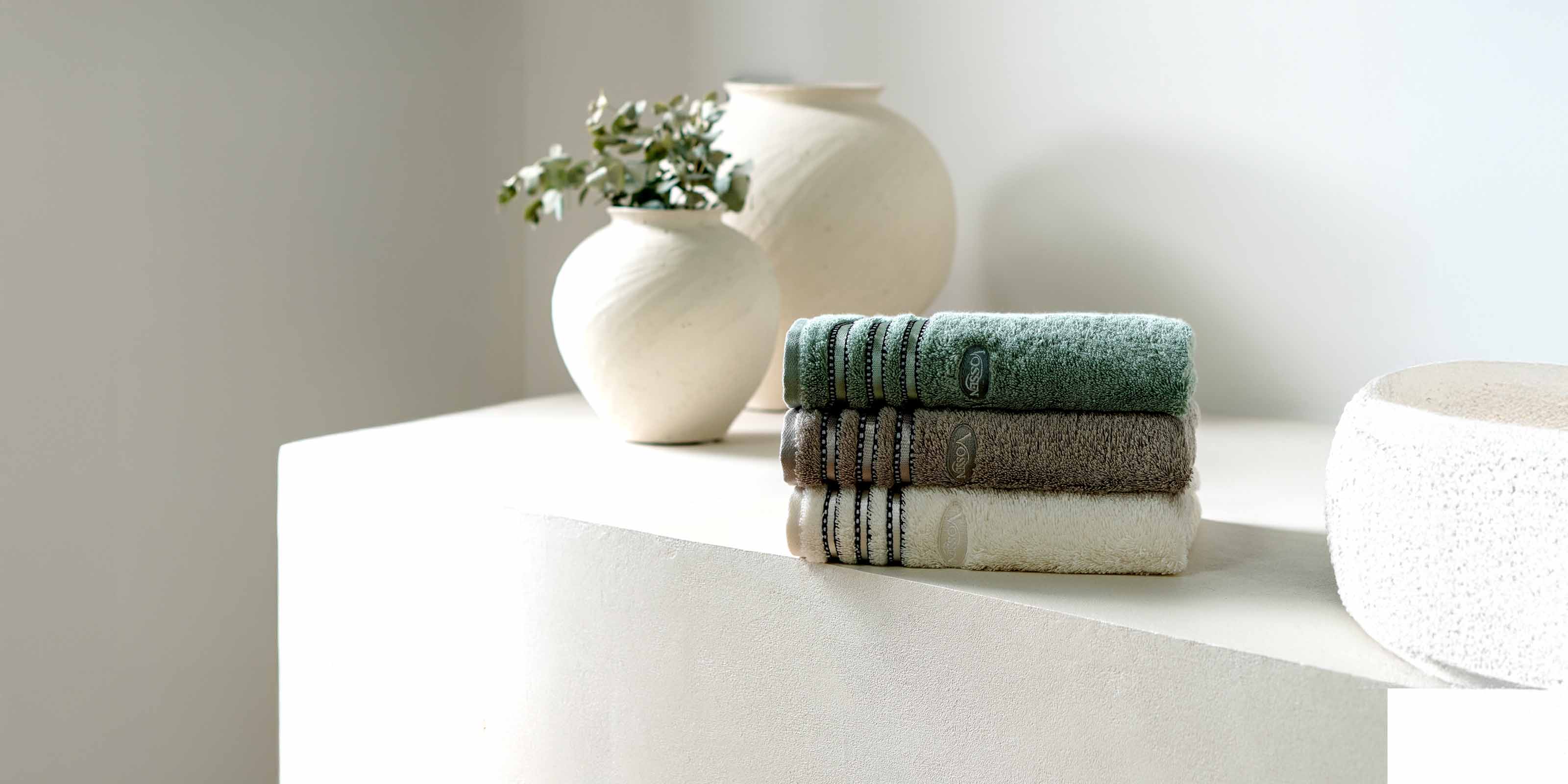 Handtuchstapel mit drei Handtüchern in Grün, Grau und Weiß liegend auf einer weißen Kommode. Dahinter stehen zwei weiße Vasen.