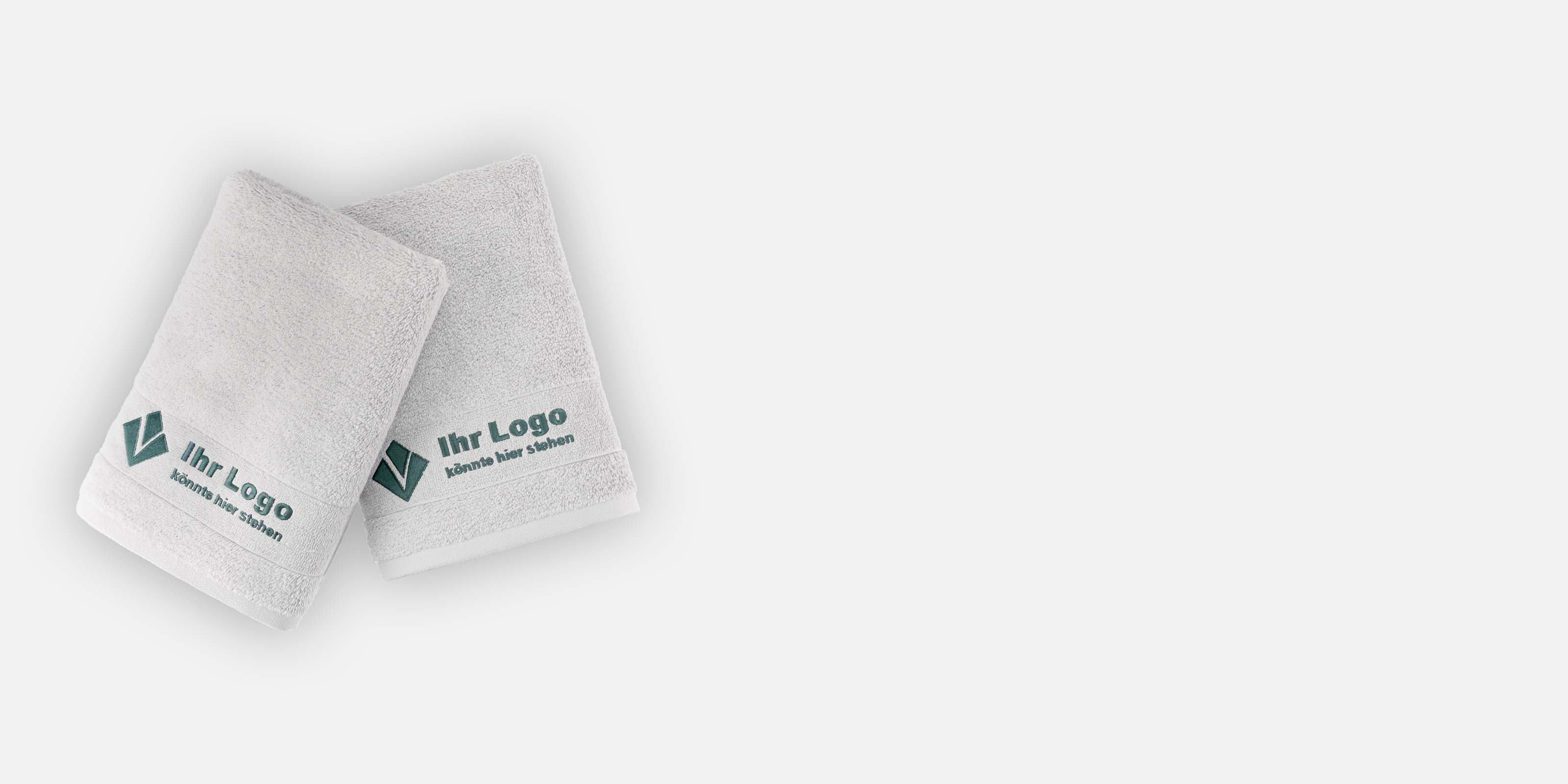 Zwei gefaltete hellgraue Handtücher liegen auf weißem Hintergrund. Darauf bestickt die Aufschrift "Ihr Logo könnte hier stehen".
