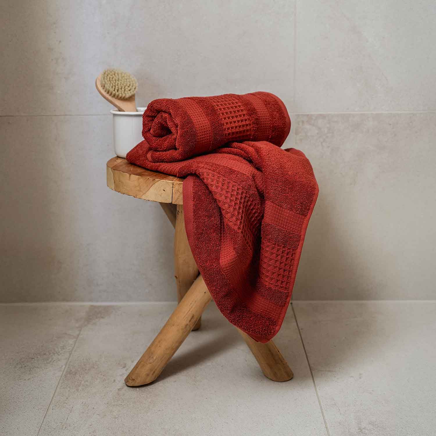 Ziegelrotes Handtuch, teils gerollt, teils ausgebreitet, liegt auf einem kleinen Hocker aus Holz. Dahinter steht eine weiße Tasse mit einer Massagebürste.