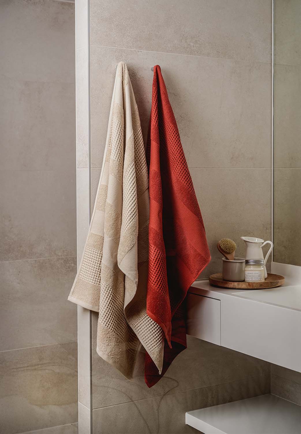 Zwei unterschiedlich gemusterte Handtücher – eines beige, eines ziegelrot – hängen nebeneinander an einer Badezimmerwand. Daneben stehen Pflegeprodukte und eine Bürste auf einem Tablett.