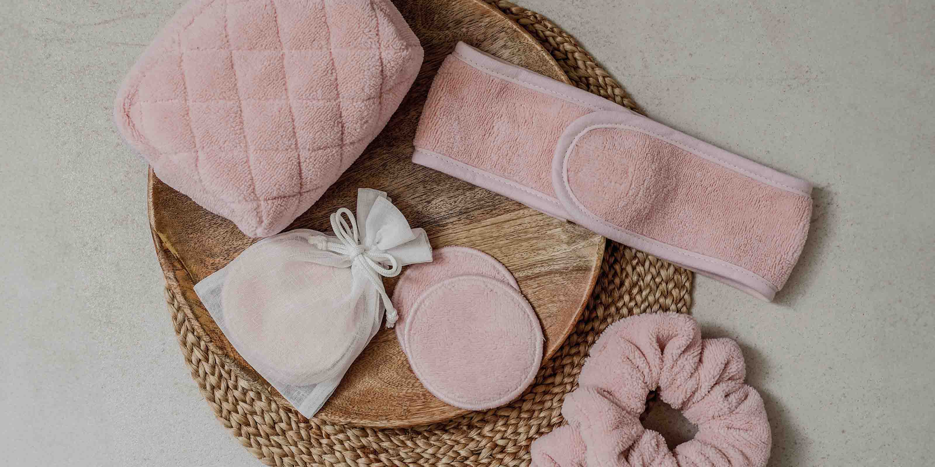 Rosa Beauty-Accessoires aus weichem Frottee – darunter ein Kosmetiktäschchen, ein Stirnband, wiederverwendbare Abschminkpads in einem Organza-Beutel und ein Haargummi – dekorativ auf einem runden Holz- und Basttablett arrangiert.