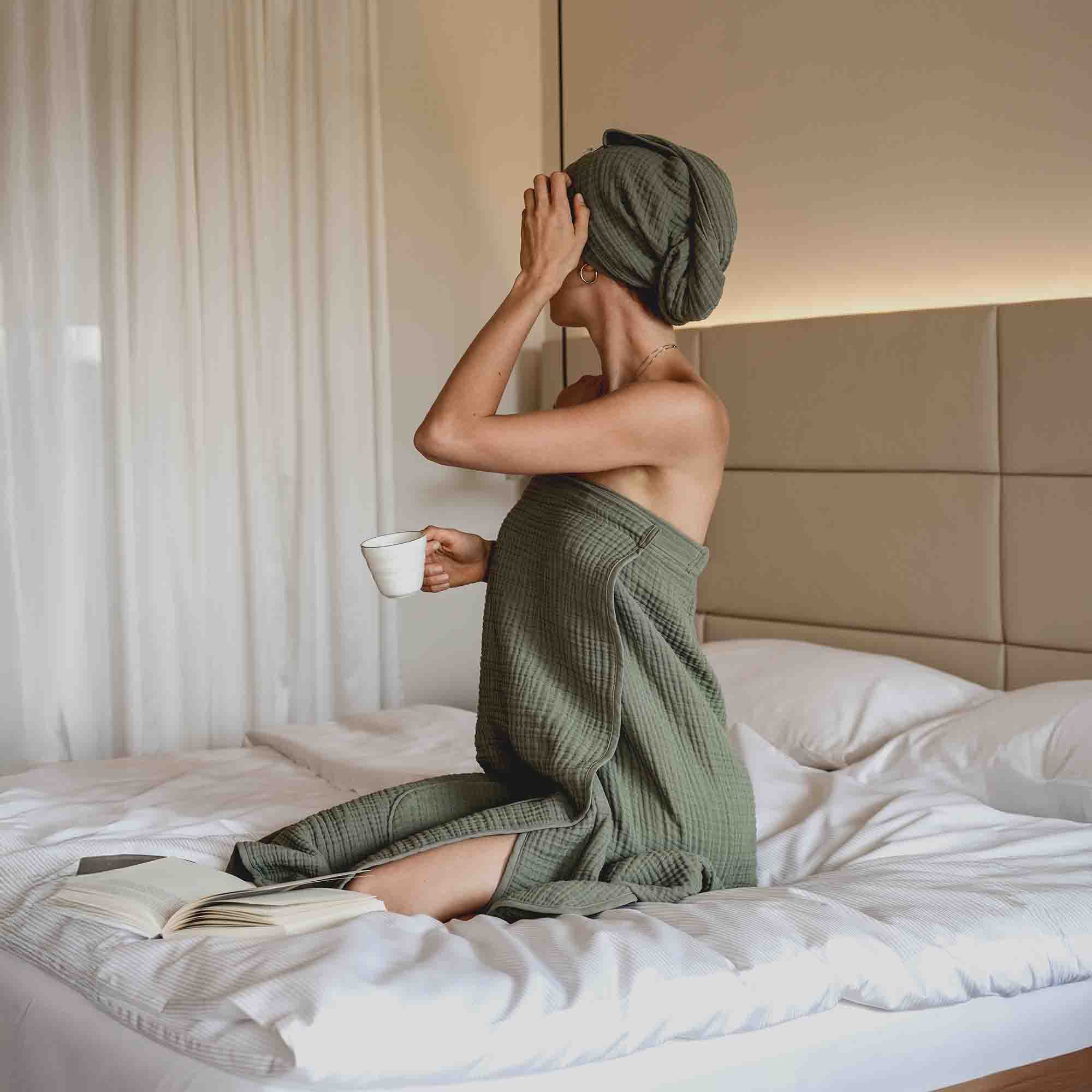 Frau in olivgrünem Handtuch mit passendem Turban sitzt auf einem Bett, hält eine Tasse in der Hand und blickt zur Seite. Neben ihr liegt ein aufgeschlagenes Buch.