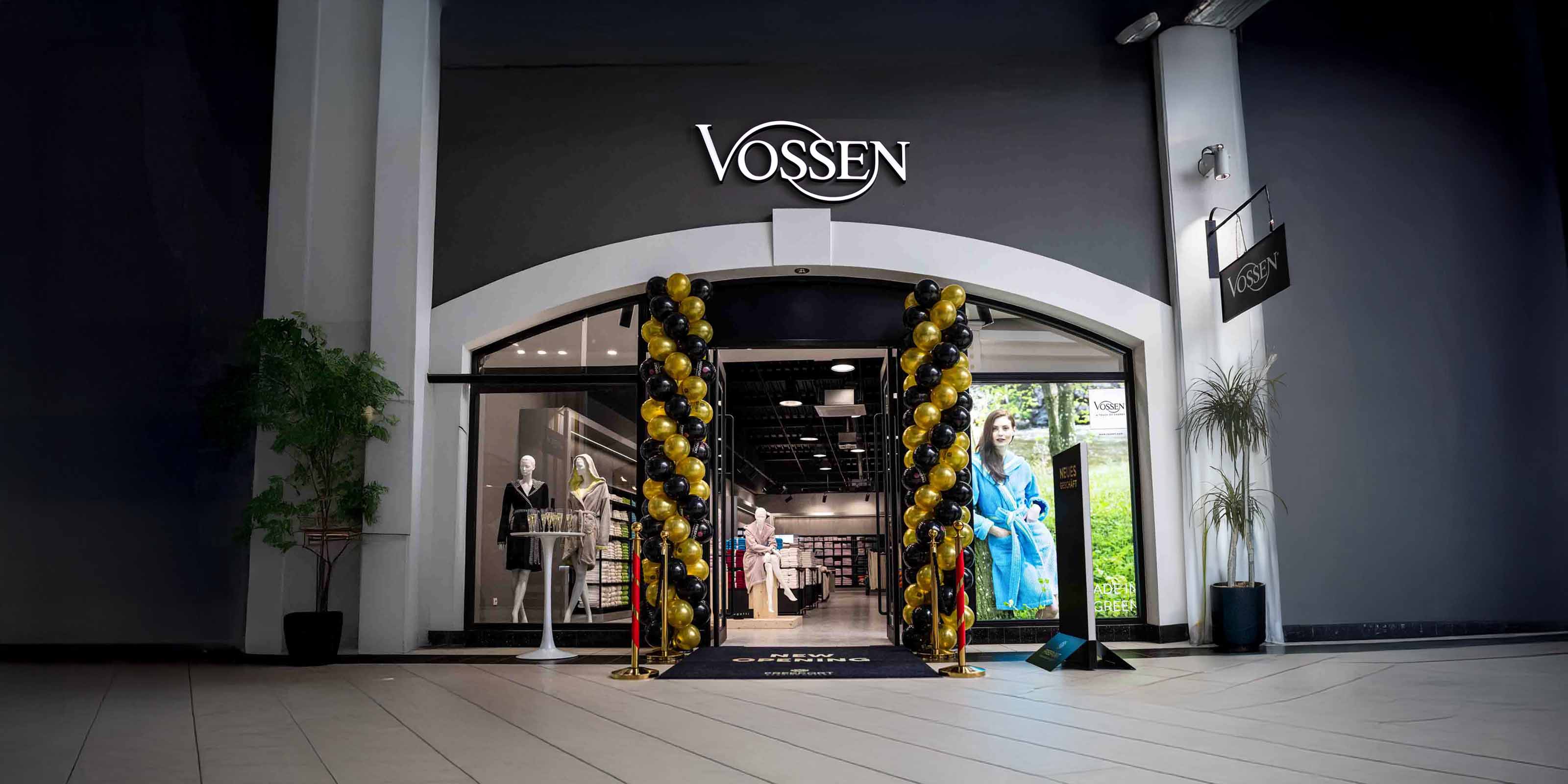 Eingangsbereich eines VOSSEN-Stores mit feierlicher Eröffnung: Schwarze und goldene Luftballons flankieren den Eingang, darüber das VOSSEN-Logo. Im Inneren sind Bademäntel und Handtücher ausgestellt.