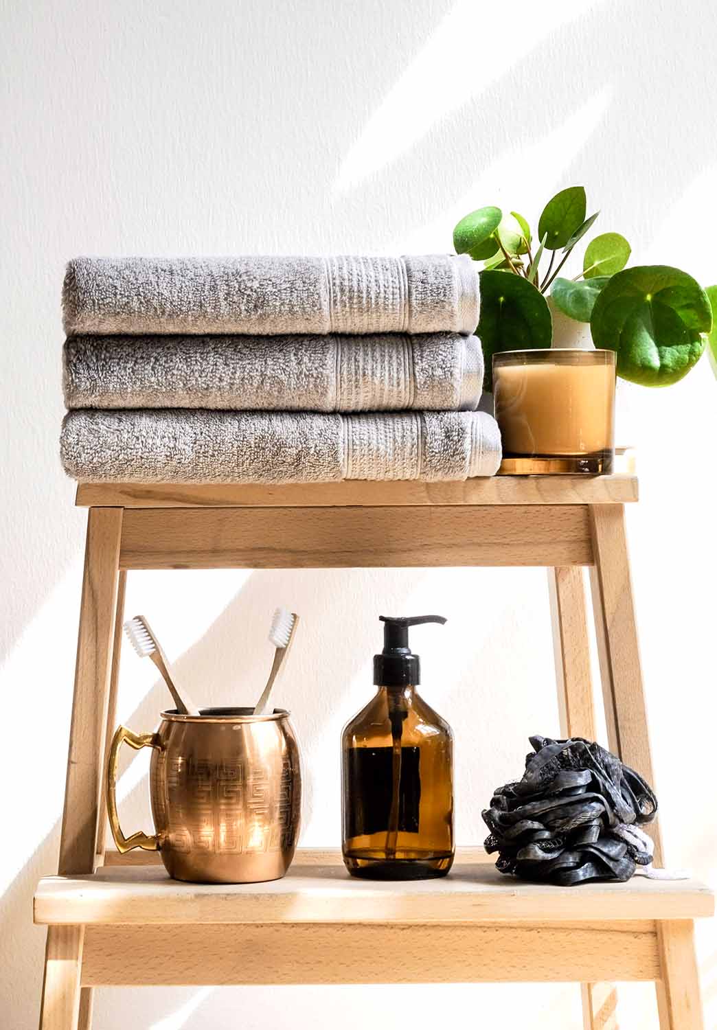 Drei graue, gefaltete Handtücher liegen ordentlich auf einem hellen Holzhocker. Daneben stehen eine kleine grüne Pflanze und ein beigefarbener Becher im Sonnenlicht.