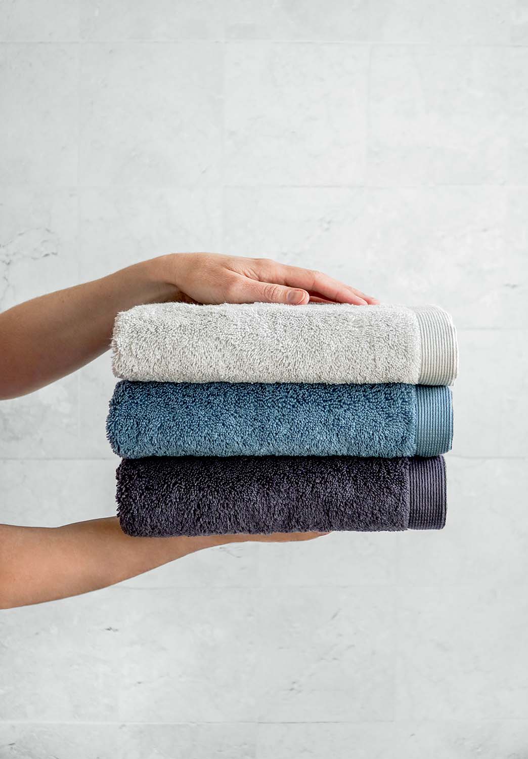 Person hält drei gefaltete Handtücher in Hellgrau, Blau und Dunkelgrau vor heller Wand.