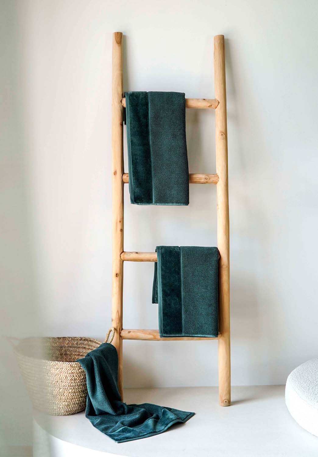 Holzleiter als Handtuchhalter mit drei dunkelgrünen Handtüchern, daneben Korb mit weiterem Handtuch, minimalistischer Raum.